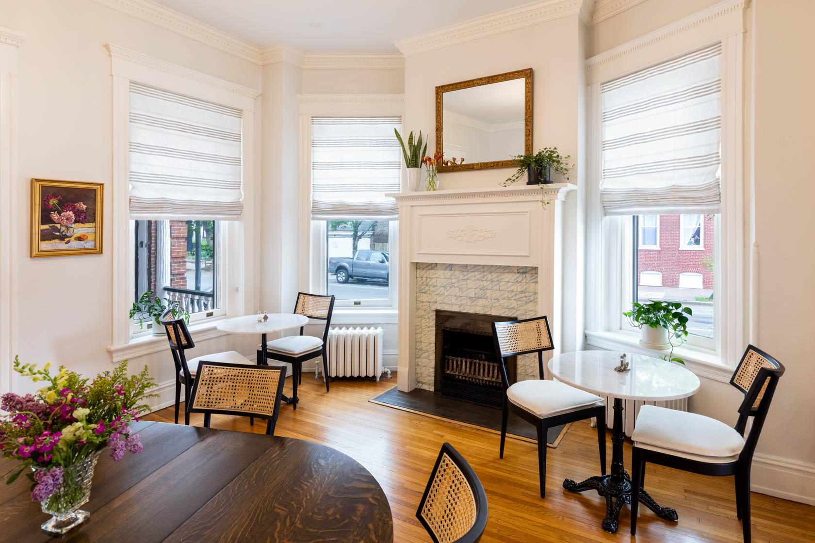 Living room with white walls, three windows with blinds, a fireplace, and round tables with chairs, decorated with plants and flowers.