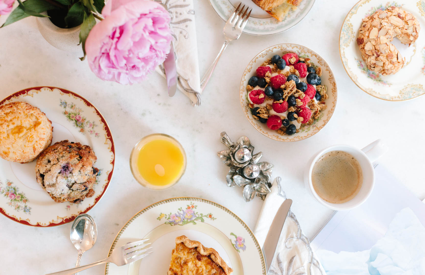 Breakfast table with pastries, berries, flowers, coffee, and juice, arranged on white tablecloth with utensils and napkins.