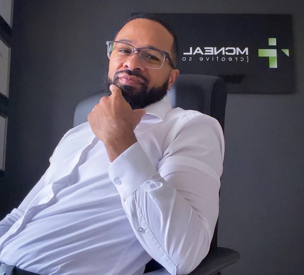 Man with glasses and beard in white shirt sitting in office chair, hand on chin, in front of a dark wall with a logo.