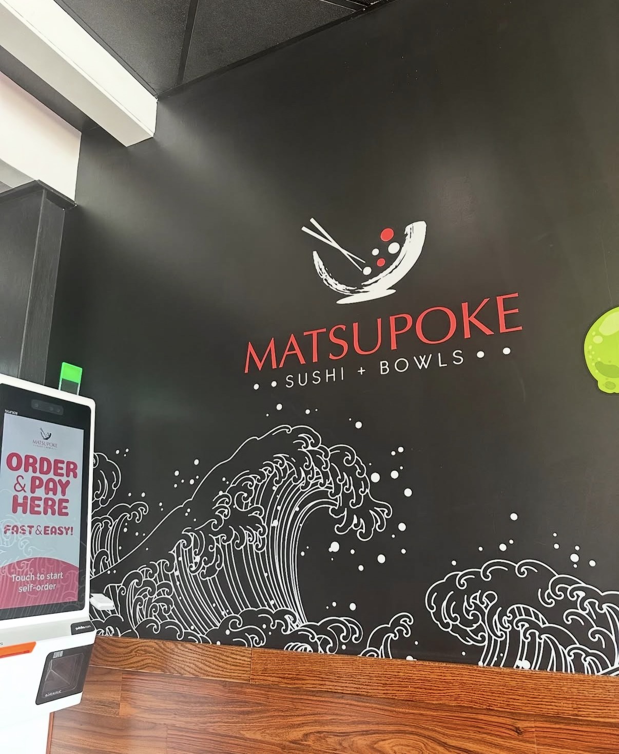 Wall with logo of sushi restaurant named MatsuPoke, featuring a bowl with chopsticks and waves below, on a black background.