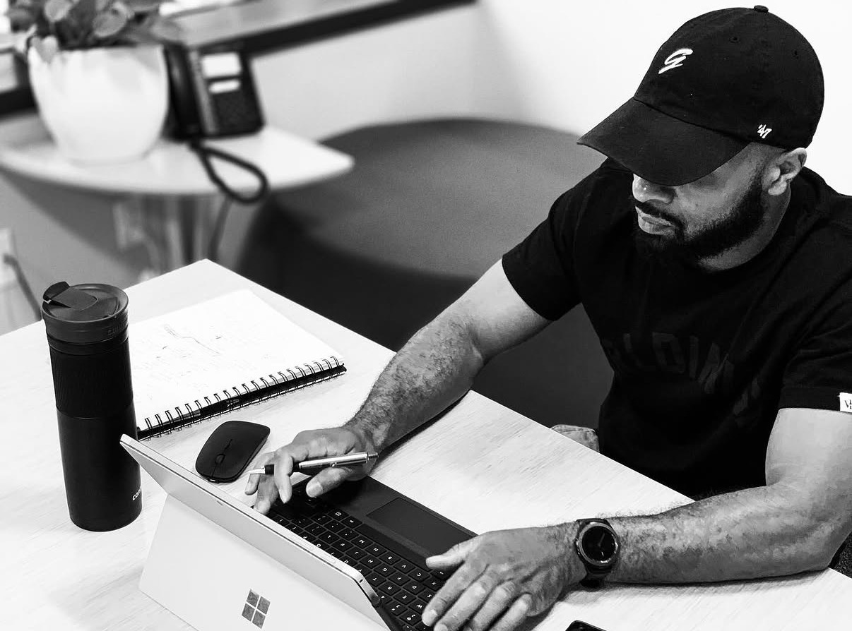 Man wearing a cap working on a laptop at a desk with a notebook, remote, and water bottle.