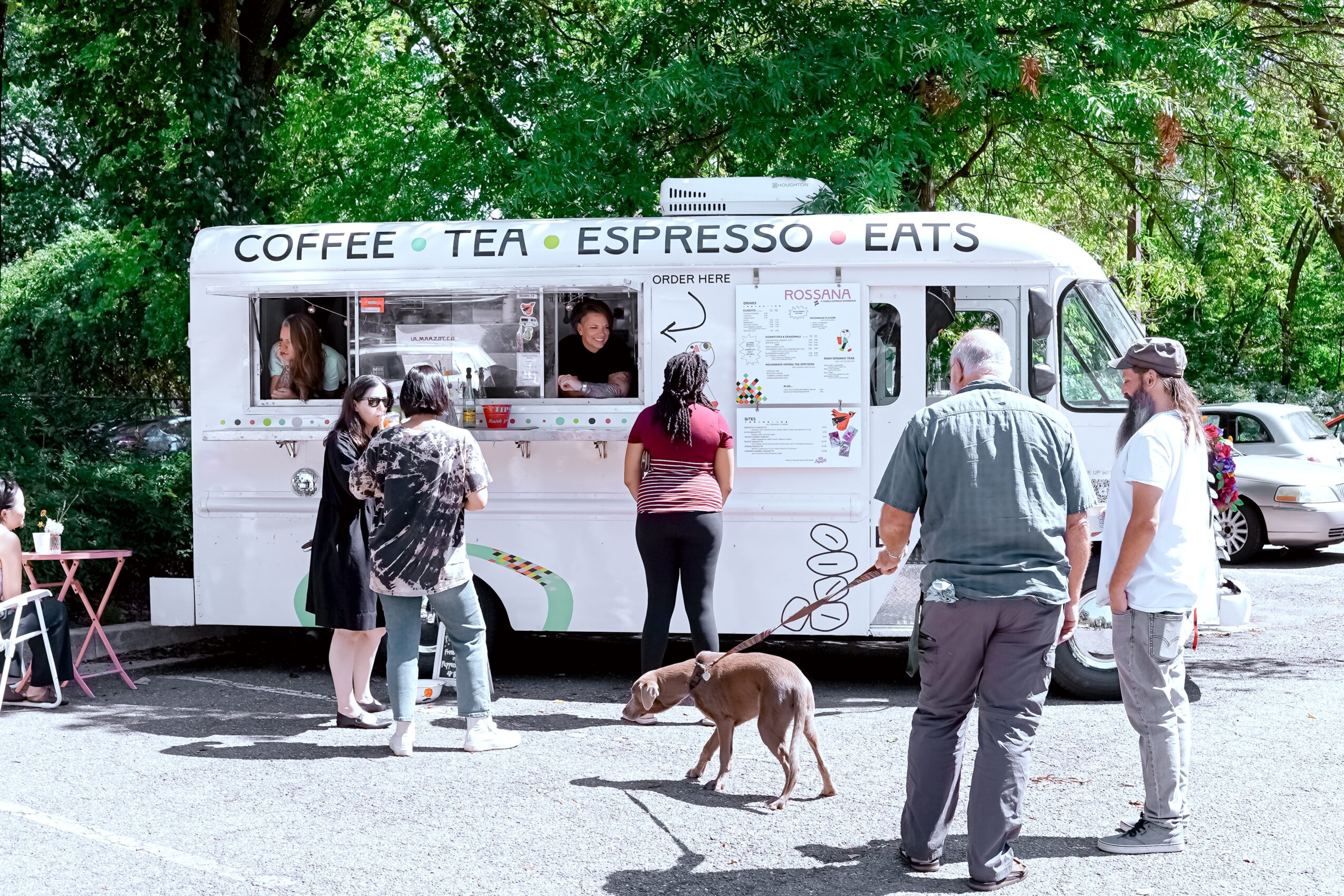 People order food at a mobile coffee stand with trees in the background, a dog on a leash, and parked cars nearby.