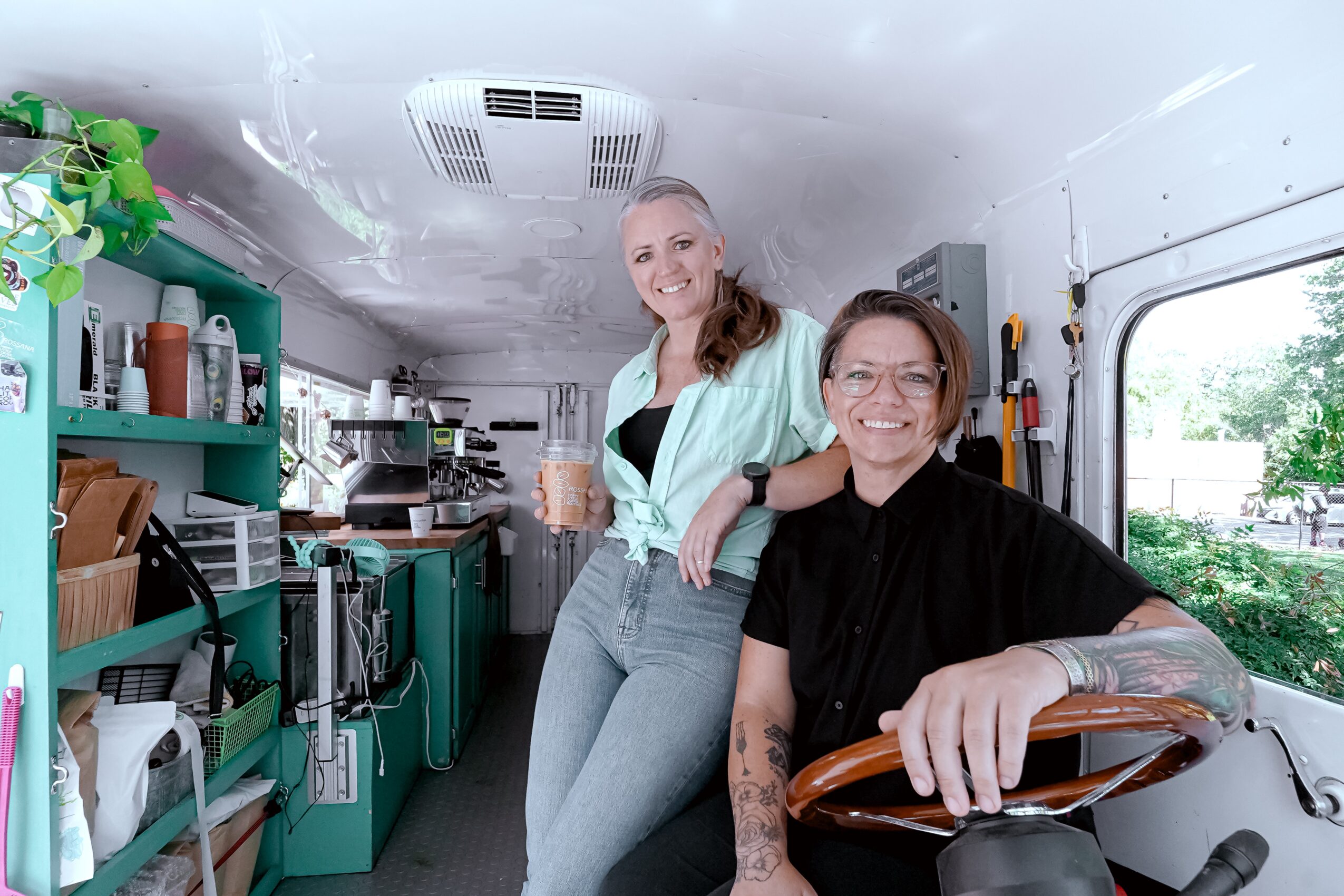 Two women inside a mobile kitchen or food truck, smiling, one holding a drink, shelves with kitchen items behind them.