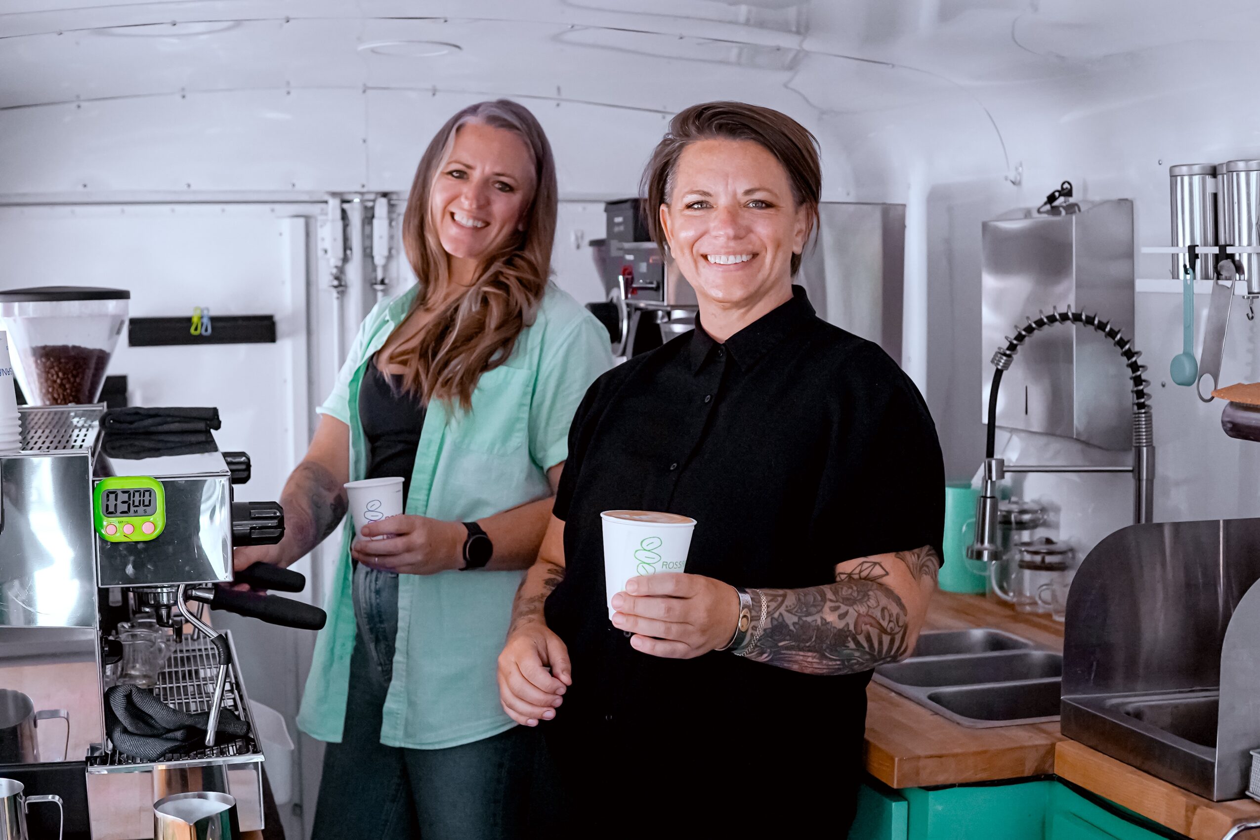 Two women smiling in a coffee shop, one holding a cup, surrounded by coffee-making equipment and a sink.