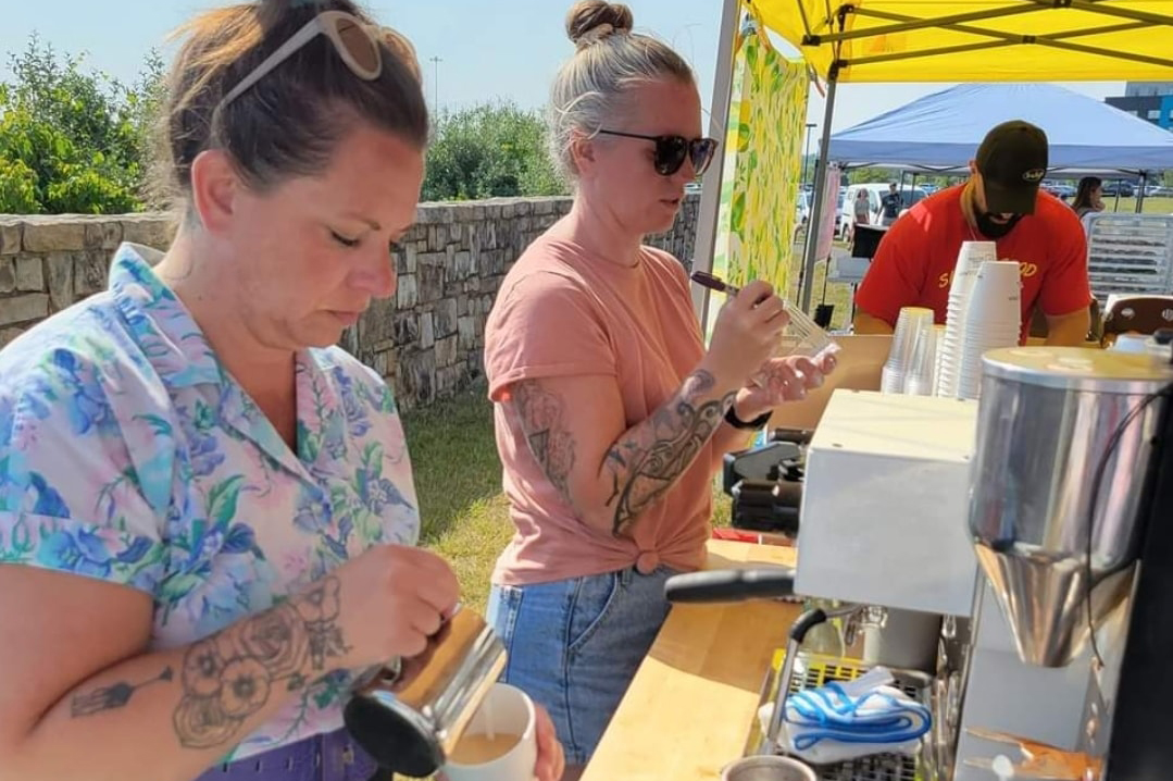 Two women at an outdoor food stall, one with tattoos and sunglasses, preparing drinks, with a man in the background under a yellow tent.