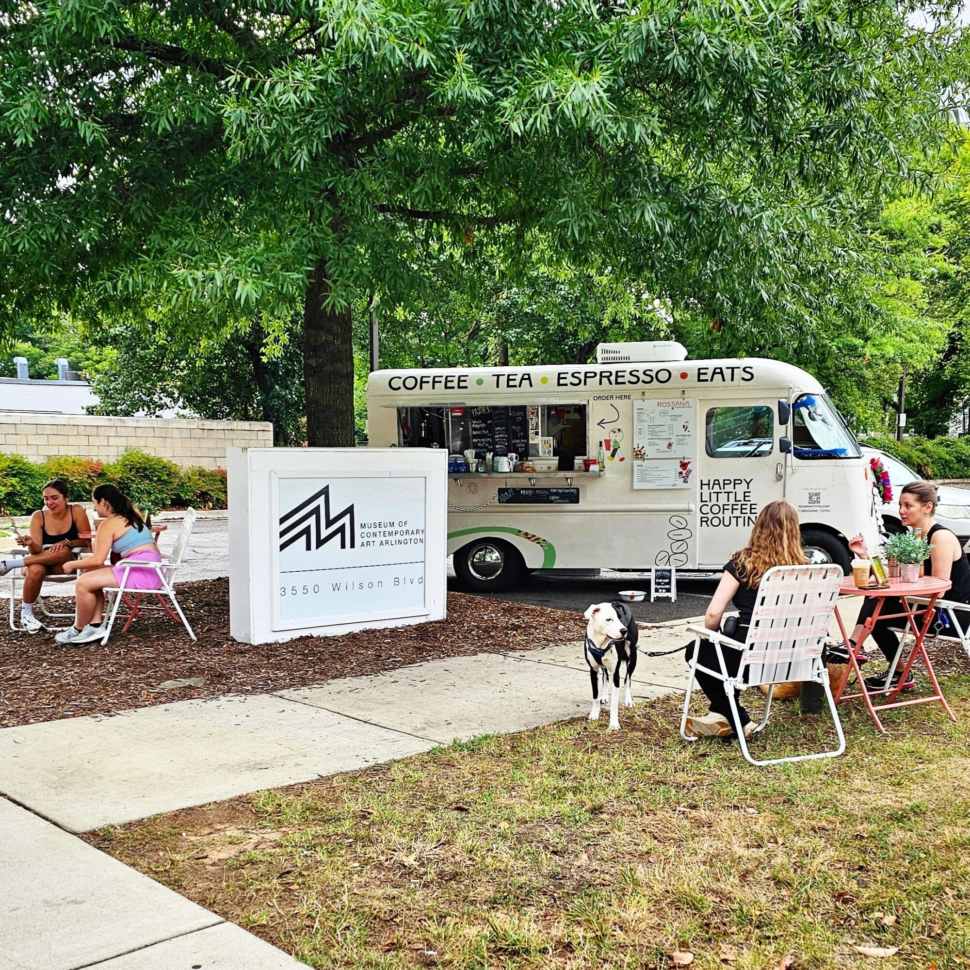 People sit at tables outside a white coffee truck with trees in the background, a dog standing nearby, and a sign in front.
