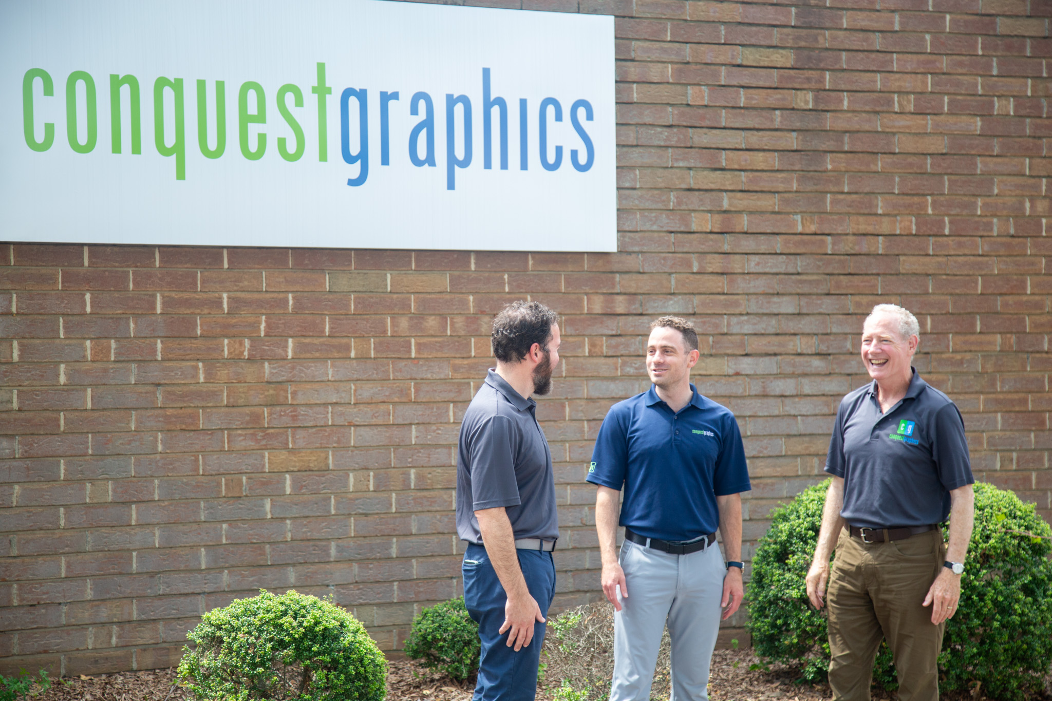 Three men standing outside near a brick wall with a sign that reads 'conquest graphics'.