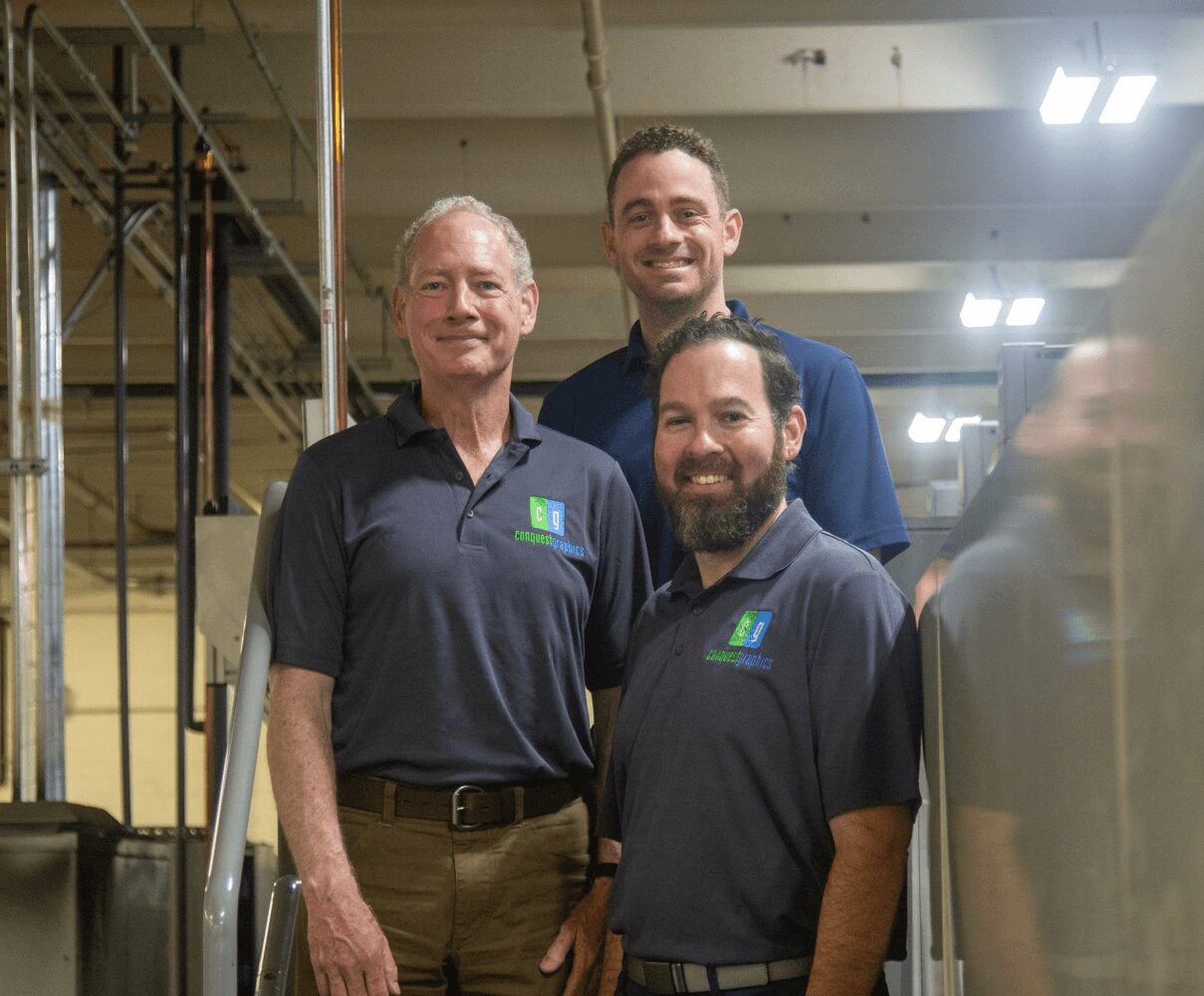 Three men standing indoors, smiling, with industrial equipment and lighting in the background.