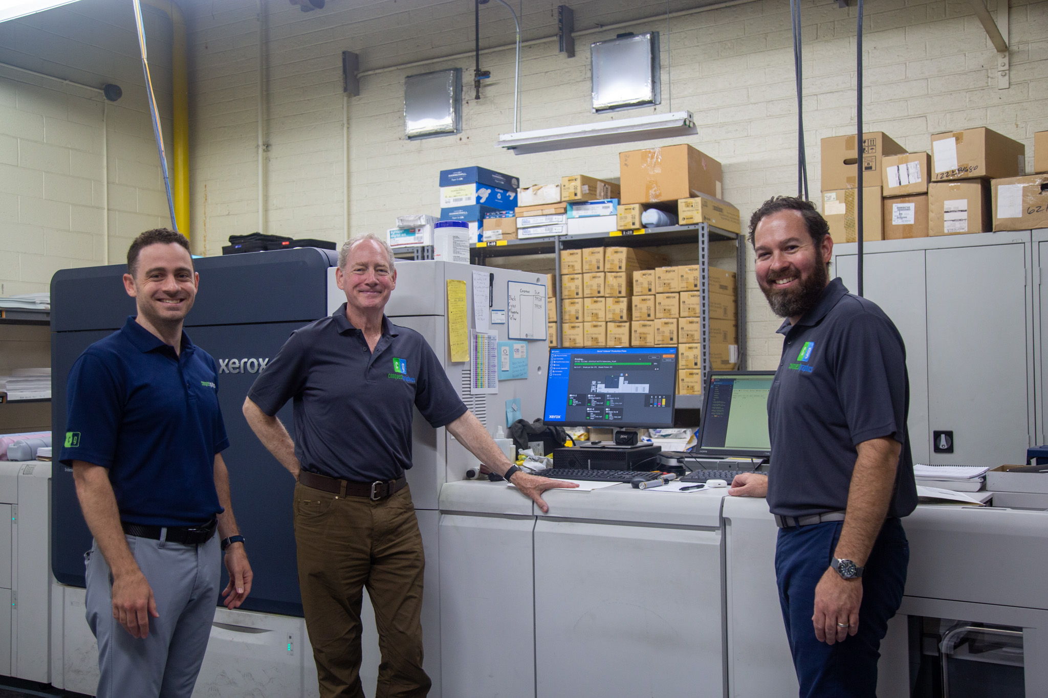 Three men standing in a warehouse with boxes and equipment behind them, smiling at the camera.