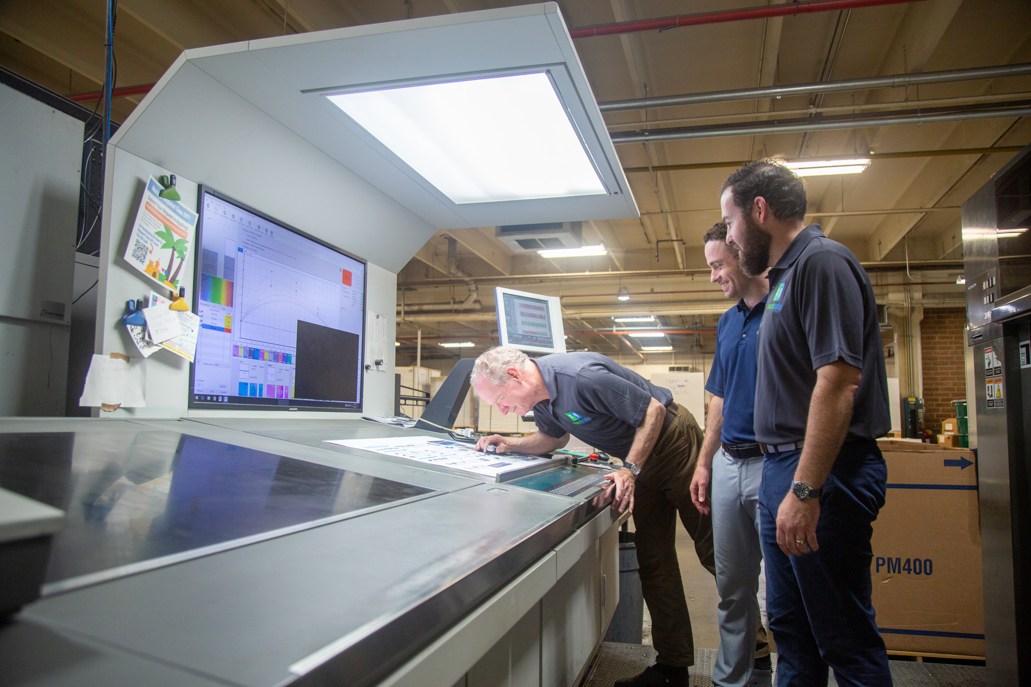 Three people examine a large screen display in a warehouse, with one person leaning over a counter, others standing nearby.
