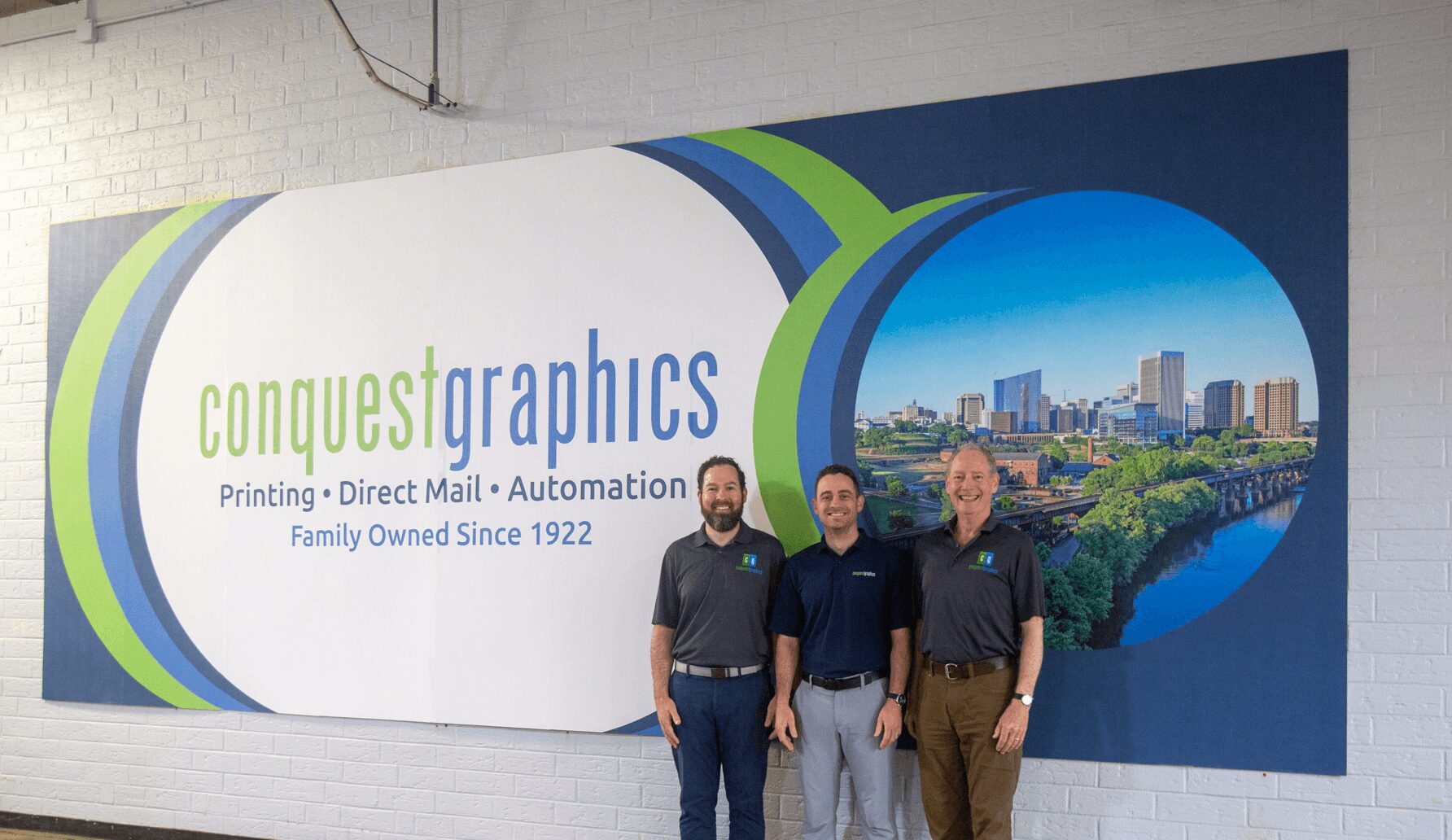 Three people stand in front of a large sign with the text 'conquest graphics' and a cityscape image, indoors.