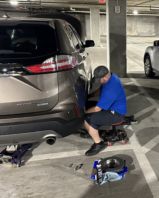 Person working on a car in an indoor parking garage, tools and equipment around them, with other cars parked nearby.