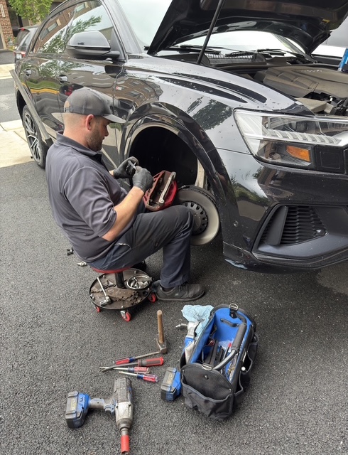 Man in gray uniform working on a black car's front wheel with tools nearby, outdoors on asphalt.