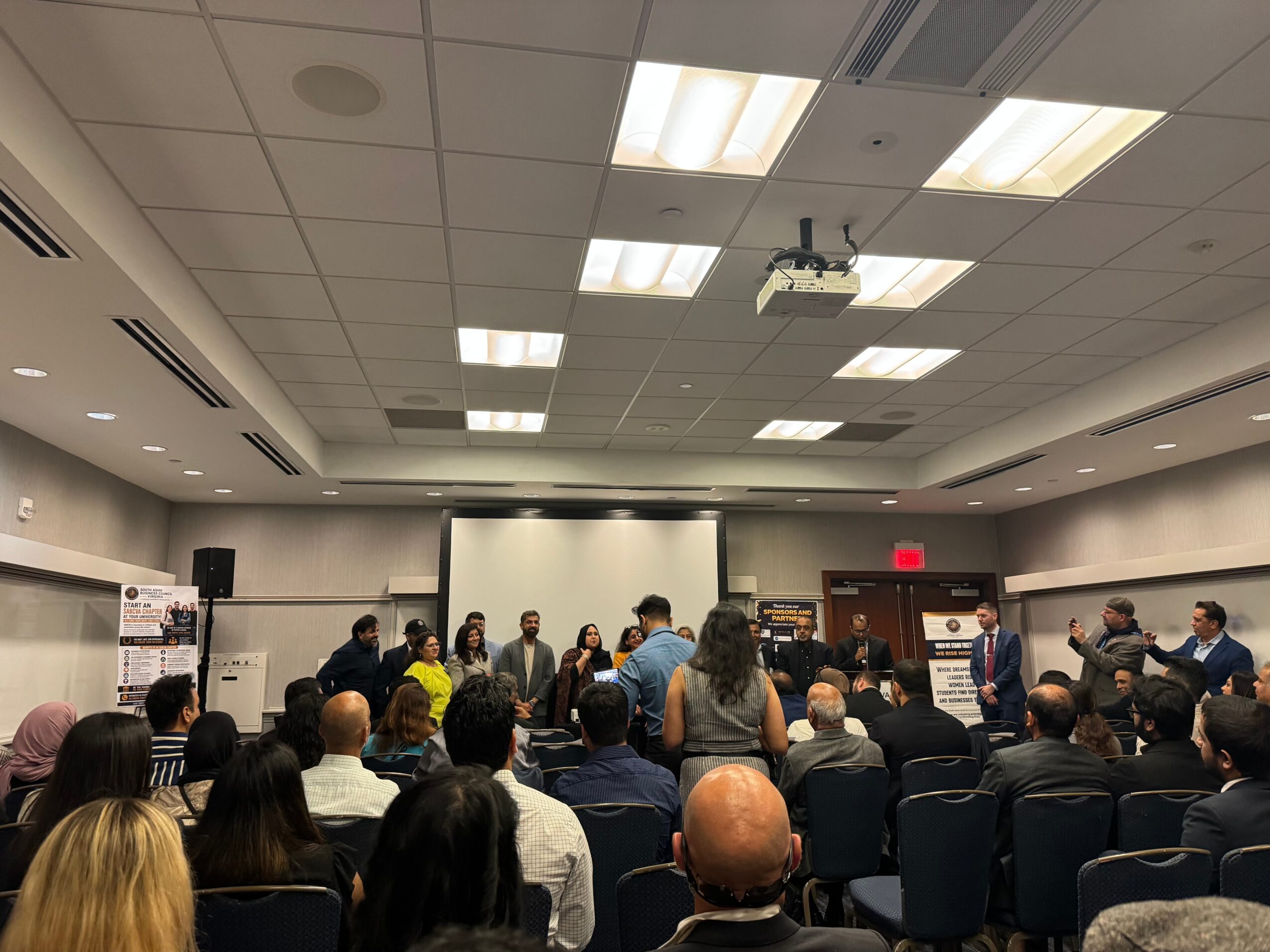 Group of people gathered in a conference room with a presentation screen at the front, audience seated and standing, ceiling lights overhead.