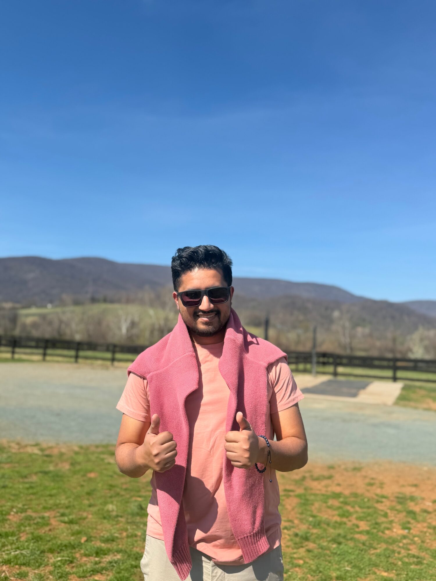 Man standing outdoors in a grassy field with mountains and a fence in the background, wearing sunglasses and a pink shirt.
