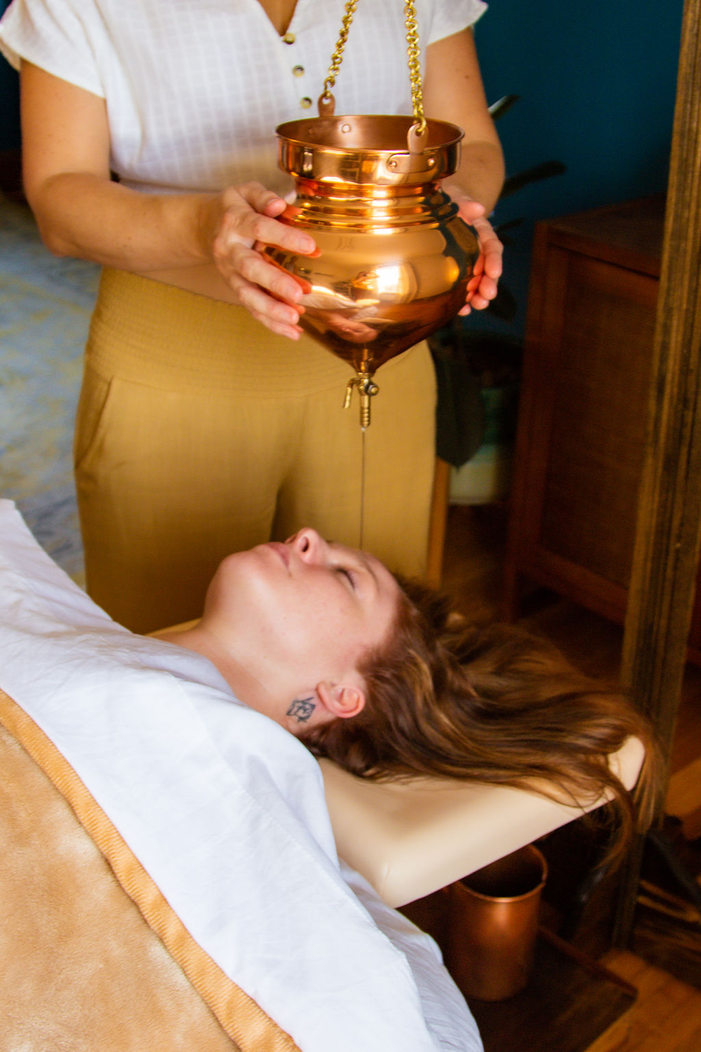 Person holding a copper urn above a woman lying on a bed, with a wooden dresser nearby.