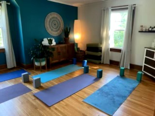Yoga mats and blocks arranged in a circle in a bright room with blue and white walls, wooden floor, and large windows.