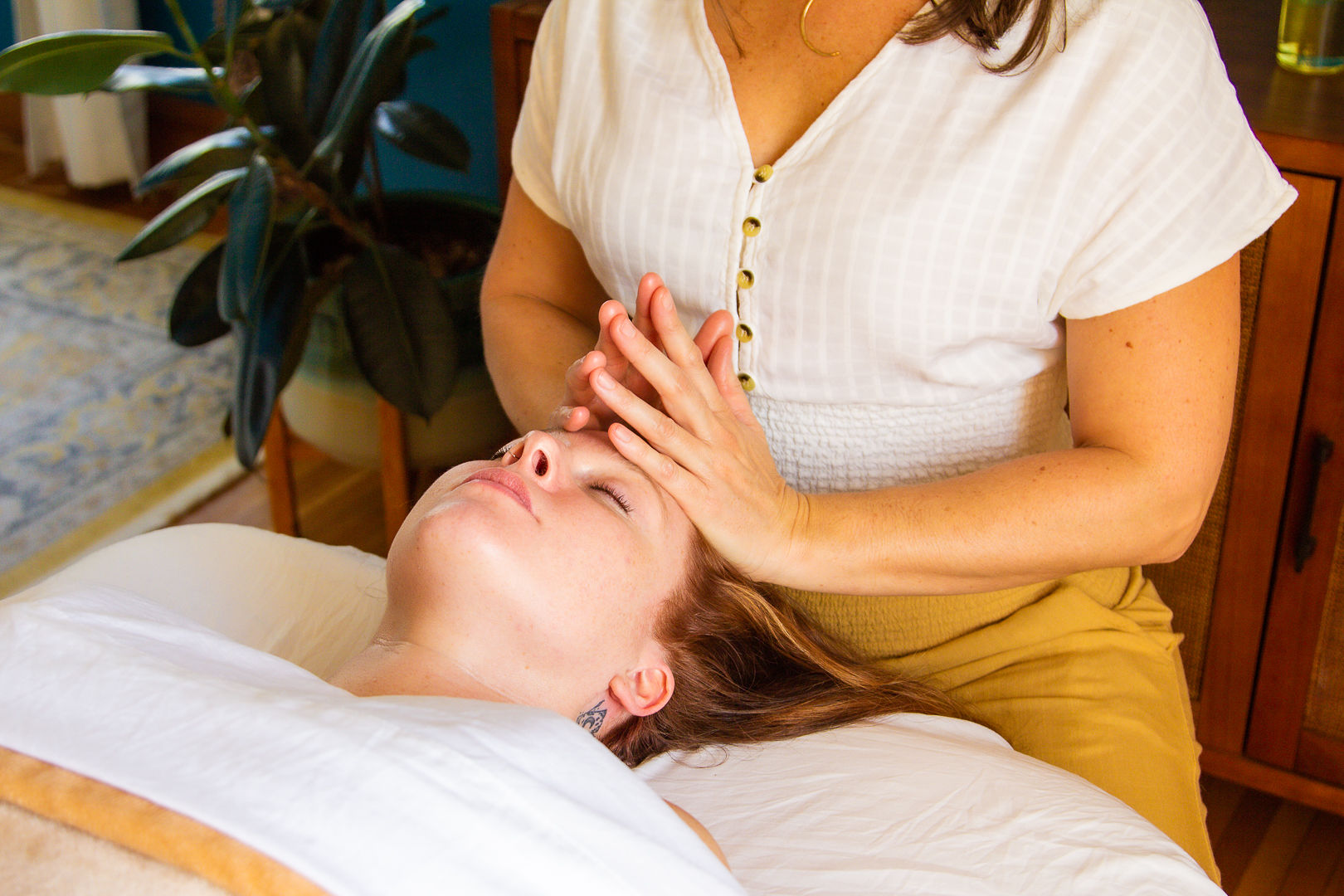 Person receiving a facial treatment from a practitioner in a cozy room with plants and wooden furniture.