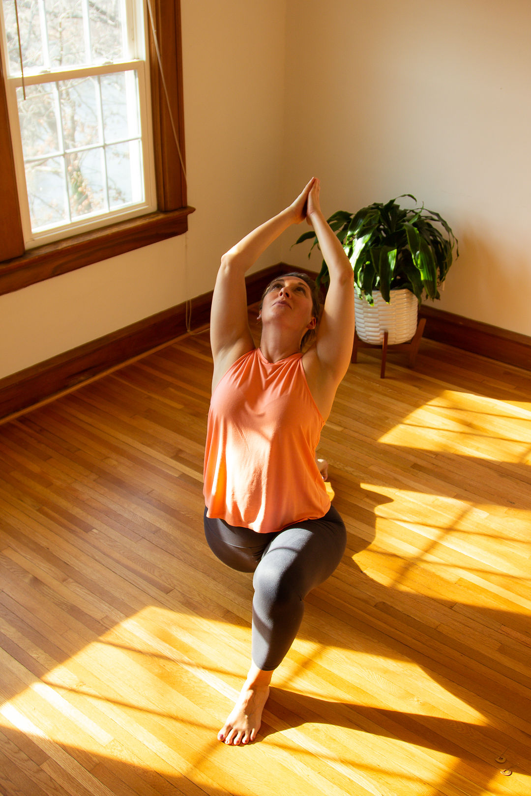 Woman practicing yoga in a sunlit room with wooden floor and a potted plant nearby.