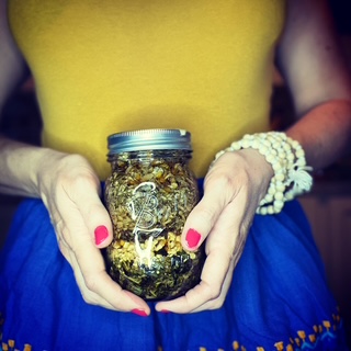 Person holding a jar of herbs or dried flowers, wearing a yellow top and blue skirt, with pink nail polish and white beaded bracelet.