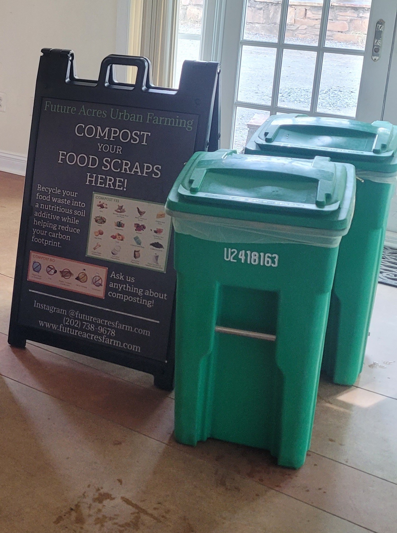 Two green compost bins next to a black signboard with white text, near a window with curtains, on a wooden floor.