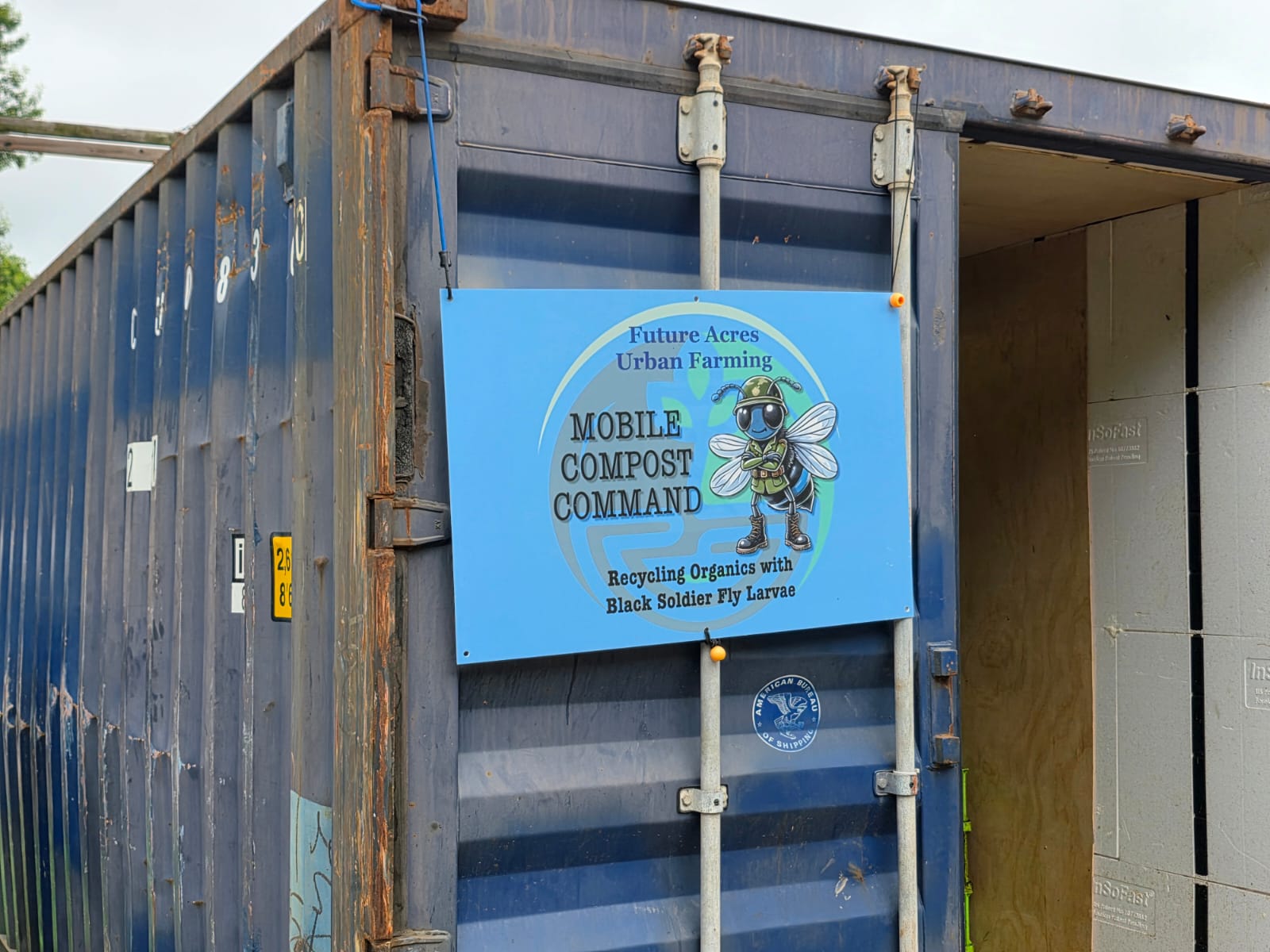 Blue sign on a container promoting urban farming, compost, and recycling with a bee illustration.