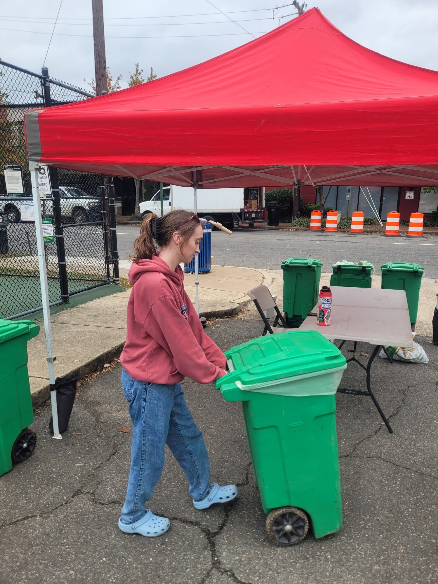 Woman in pink jacket and jeans standing near green trash bin under red canopy, with tables and chairs nearby.