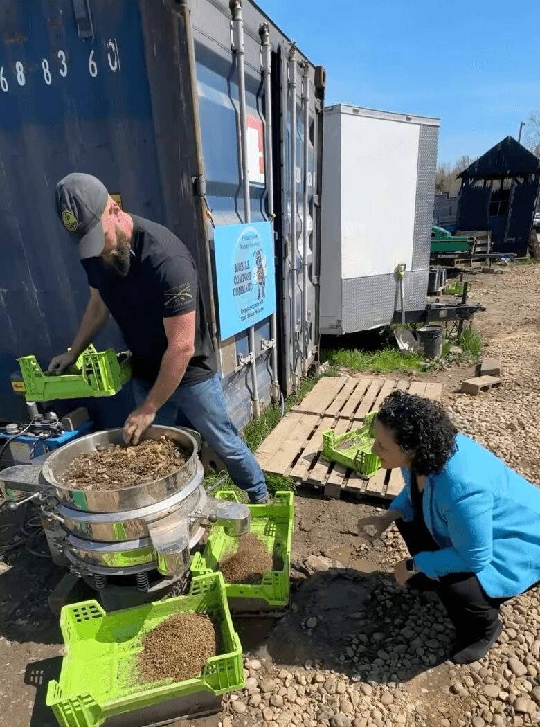Man and woman working outdoors with green crates and a large metal container, near a black container and wooden pallets.