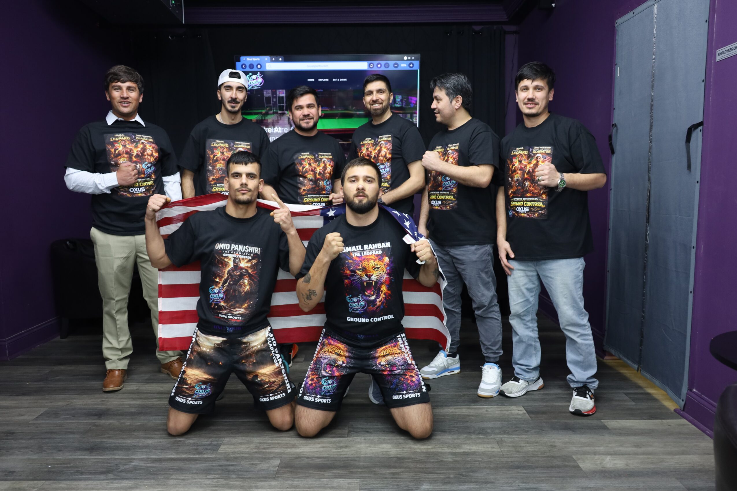 Group of nine people in a room, some holding trophies, wearing matching black T-shirts with colorful designs, standing and kneeling in front of a table with an American flag-themed cloth.