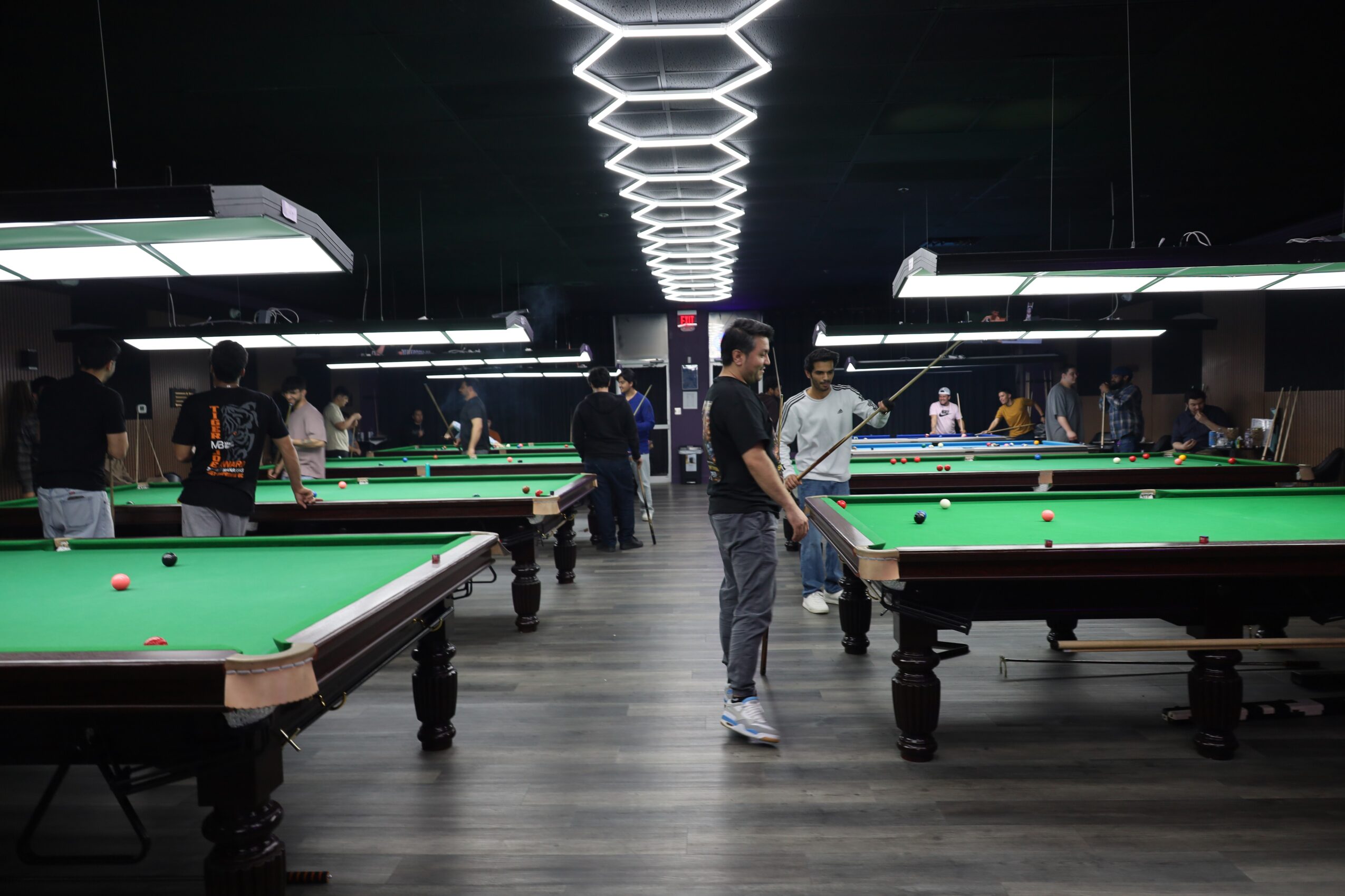 People playing pool in a modern, dimly lit billiards hall with bright ceiling lights and multiple tables.