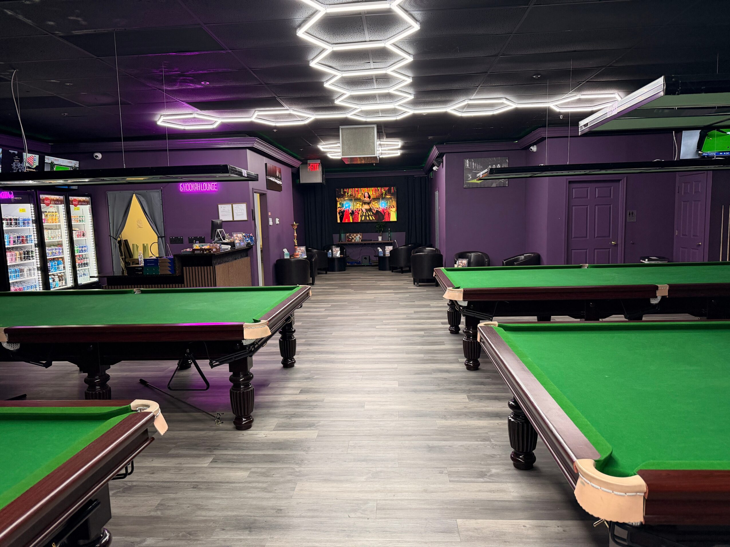 Pool hall with four green tables, purple walls, vending machines, and modern ceiling lights, empty of people.
