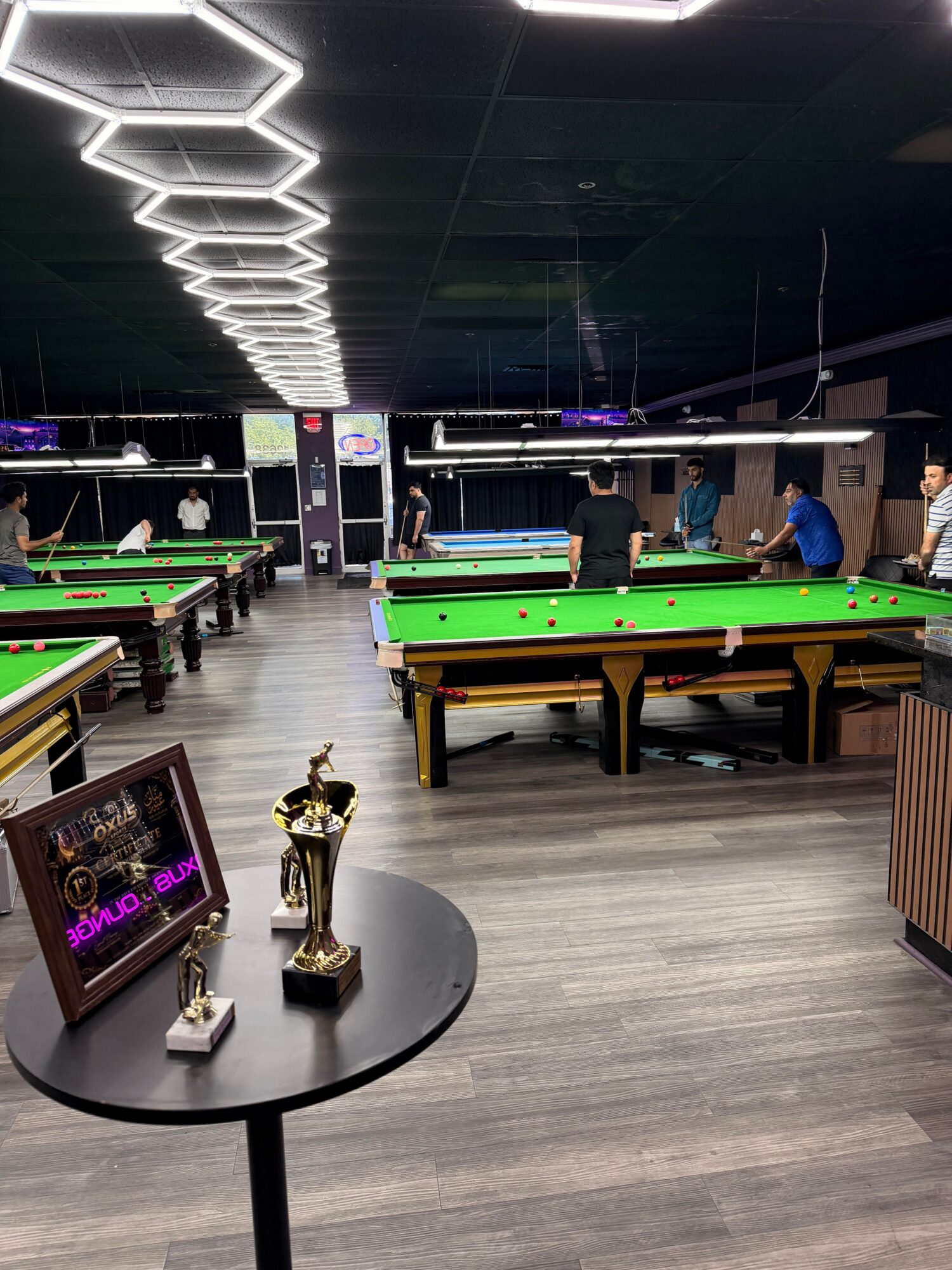 Pool hall with multiple green pool tables, people playing, and trophies on a round table in the foreground.