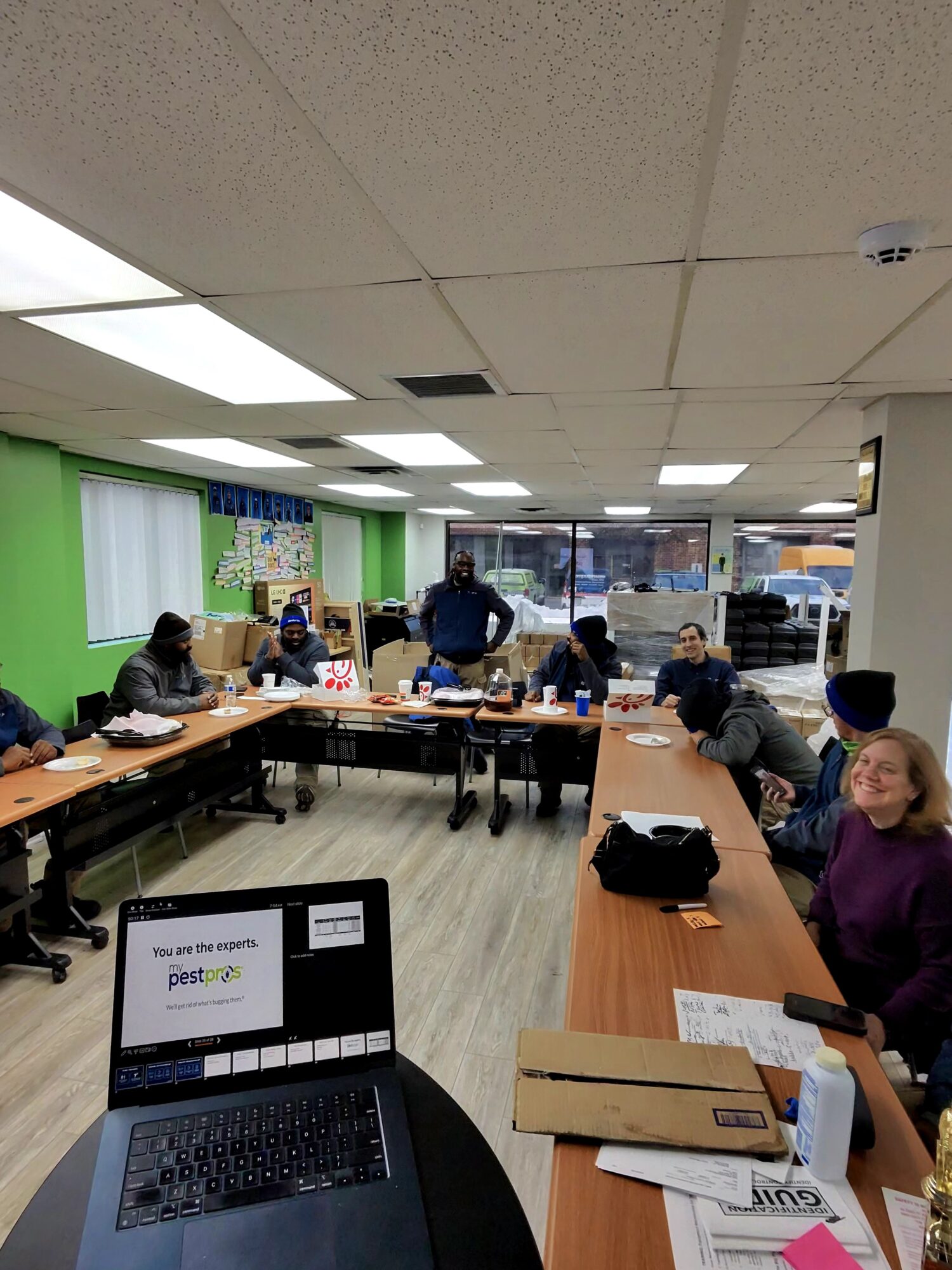 People seated around tables in a room with green and white walls, some working on laptops and others talking.