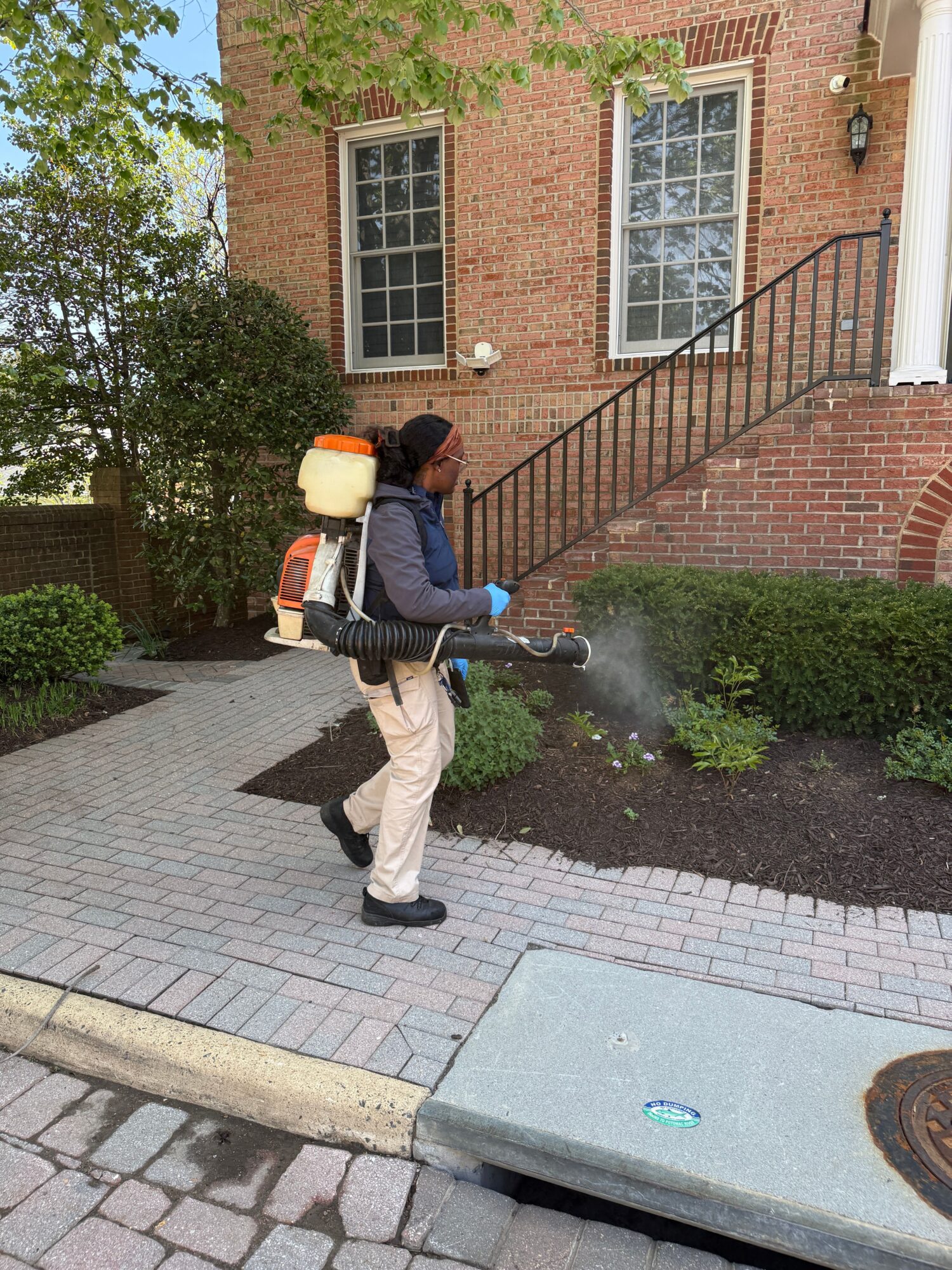 Person wearing protective gear sprays disinfectant on sidewalk near building with brick walls and windows.