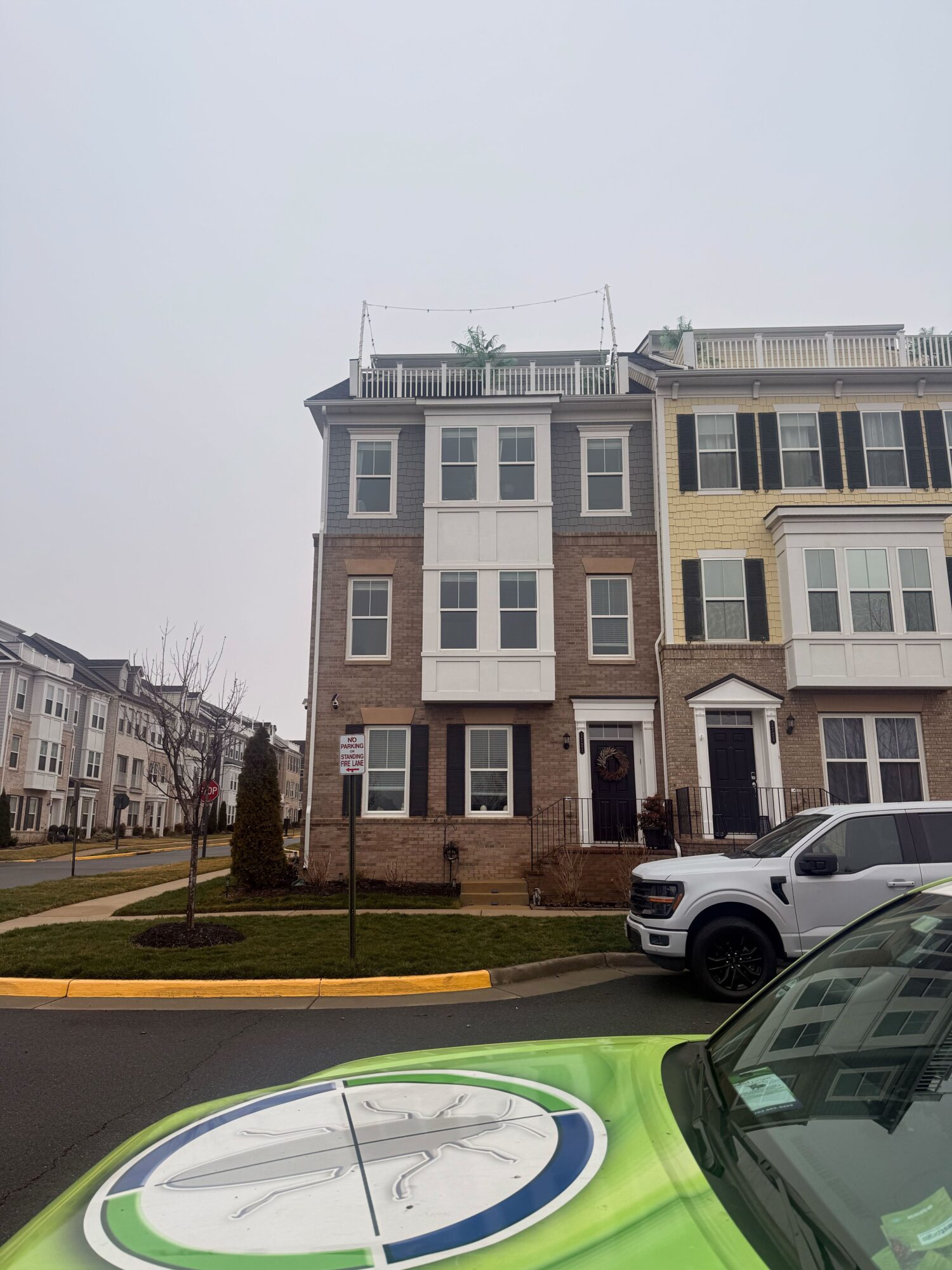 Multi-story residential building with a white car parked in front, overcast sky, and a small landscaped area with a tree.