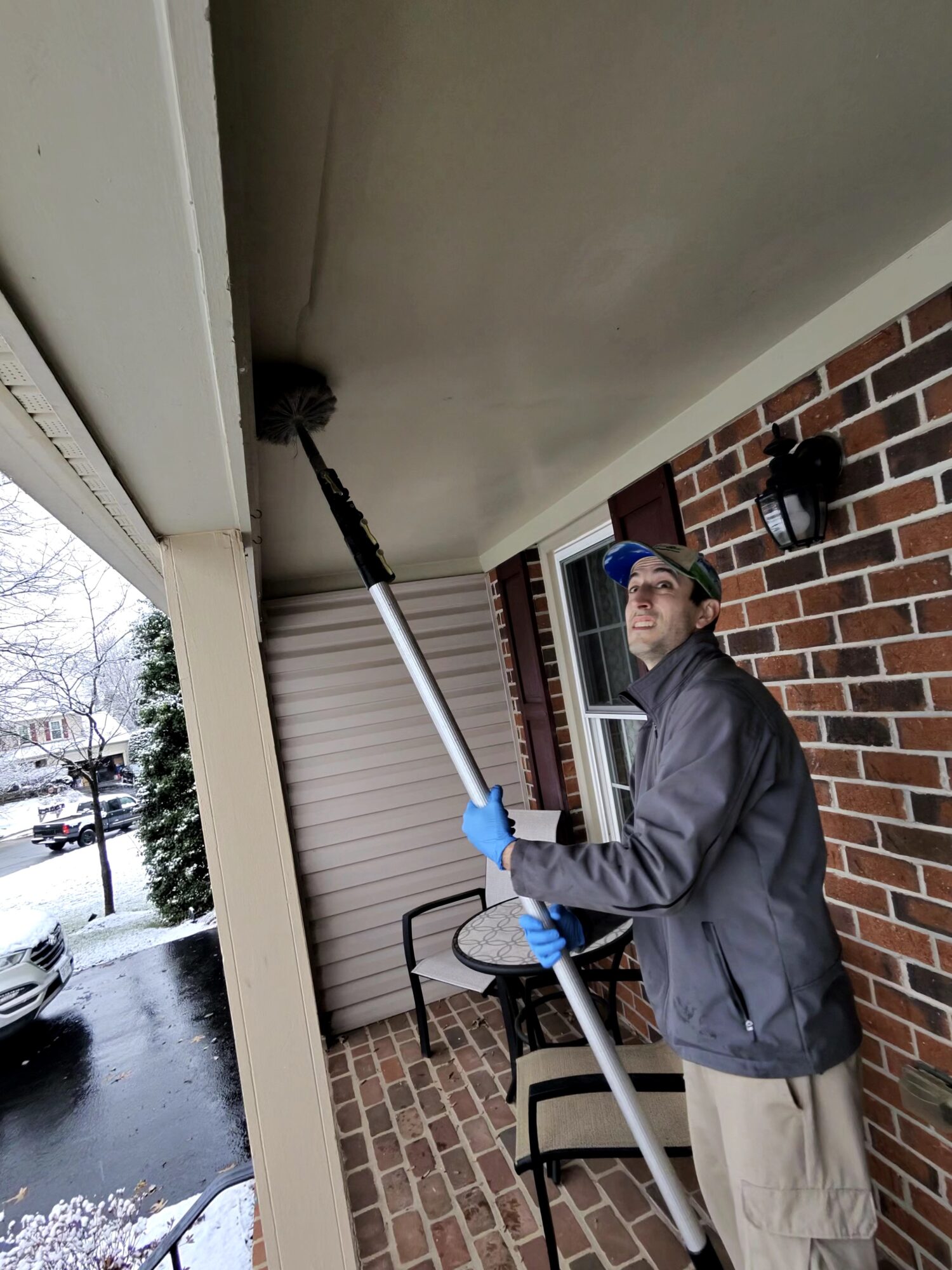 Person cleaning ceiling with extendable pole outside house, brick wall background, winter scene with snow, person wearing glasses and jacket.