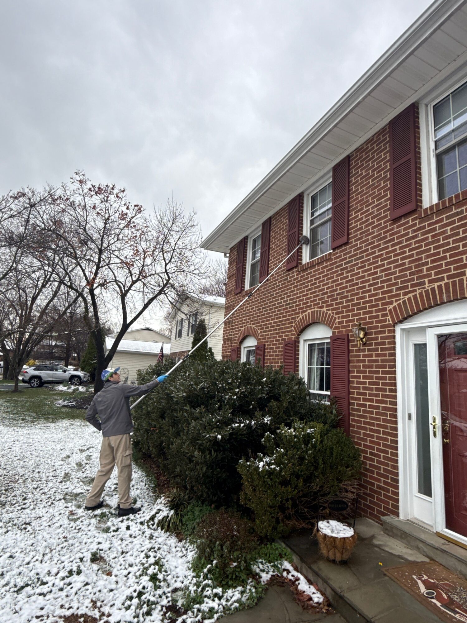 Person standing outside near a brick building with snow on the ground and a large bush, overcast sky.