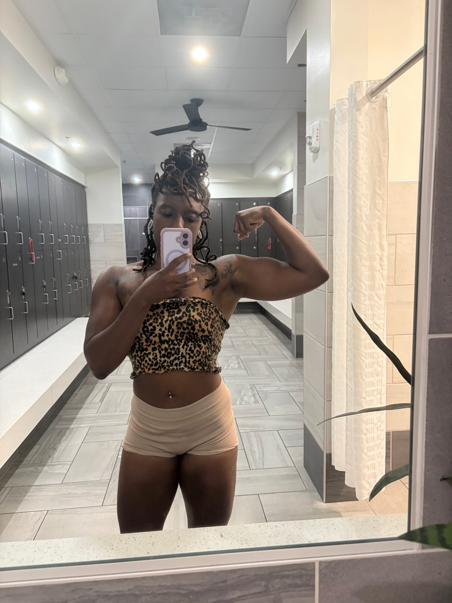 Person taking a mirror selfie in a room with white walls, ceiling fan, and tiled floor, wearing a leopard print top and beige shorts.