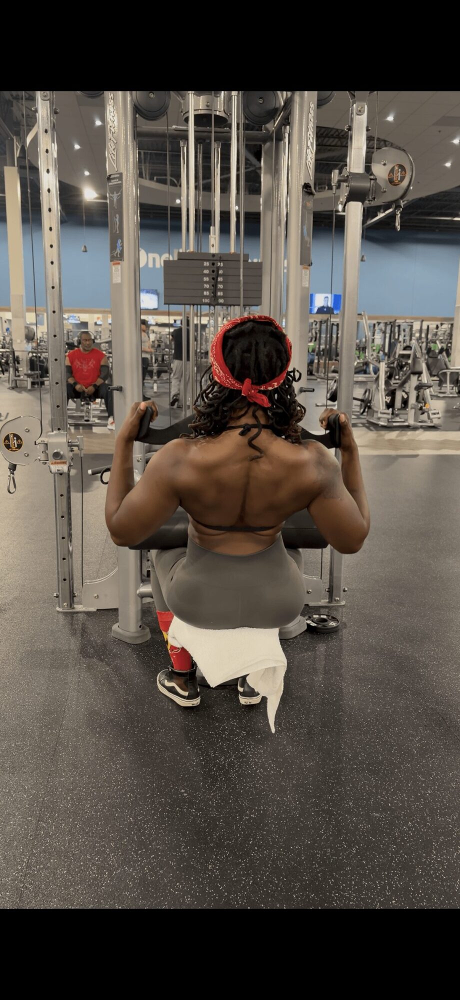 Person kneeling in gym, facing away, flexing arms, with gym equipment in background.