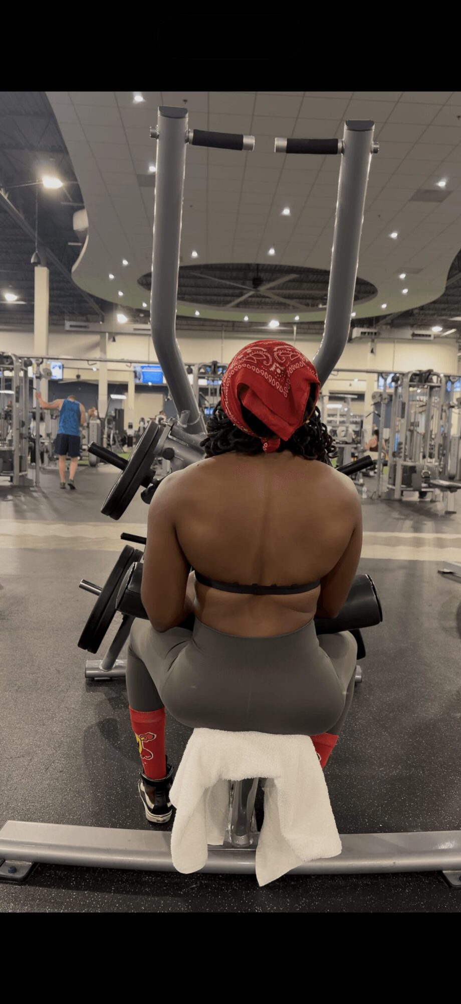 Person sitting on a workout bench in a gym, facing away, wearing a red headscarf and black sports bra.