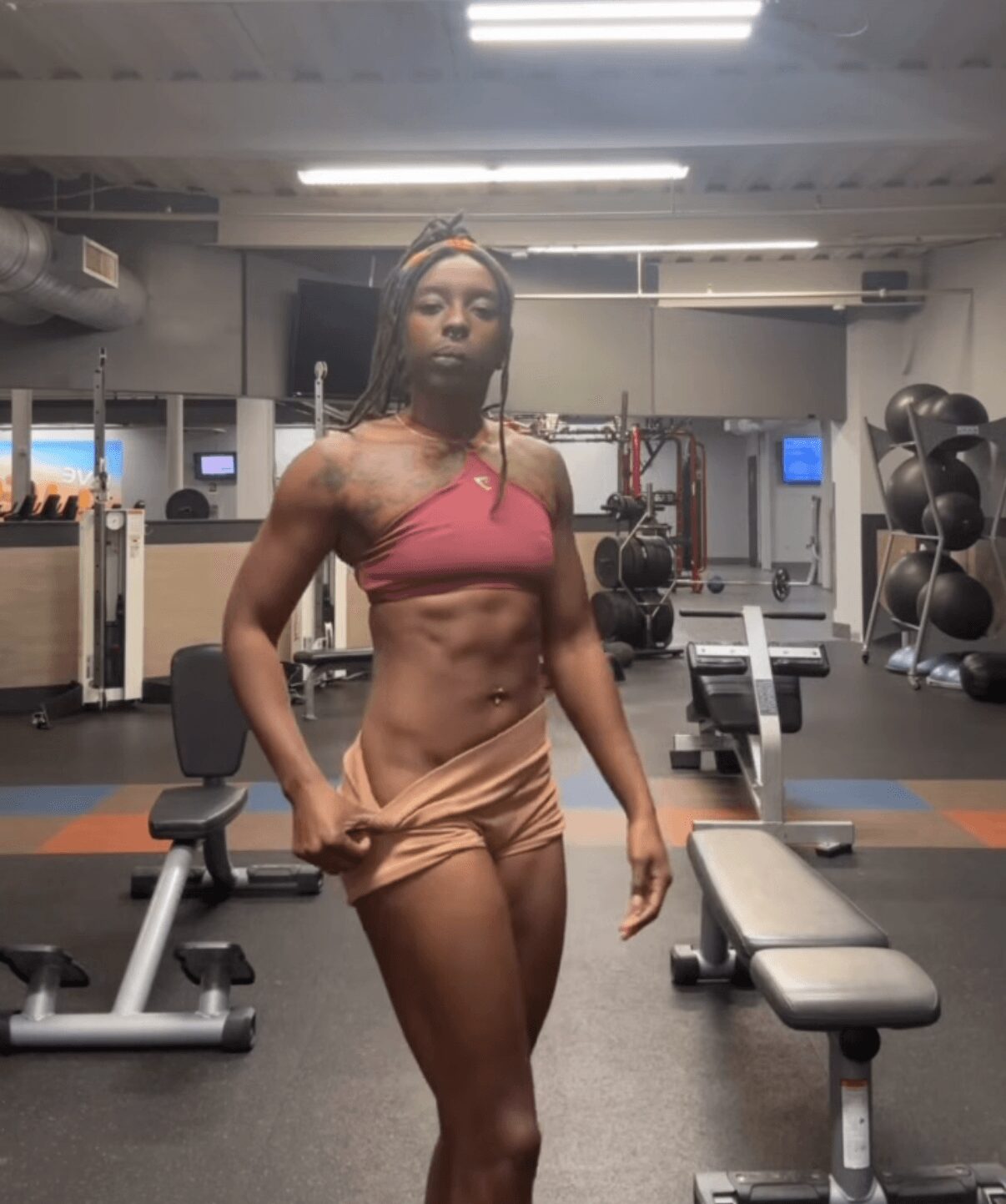 A woman with a toned physique in workout clothes stands in a gym, holding a dumbbell, with gym equipment in background.