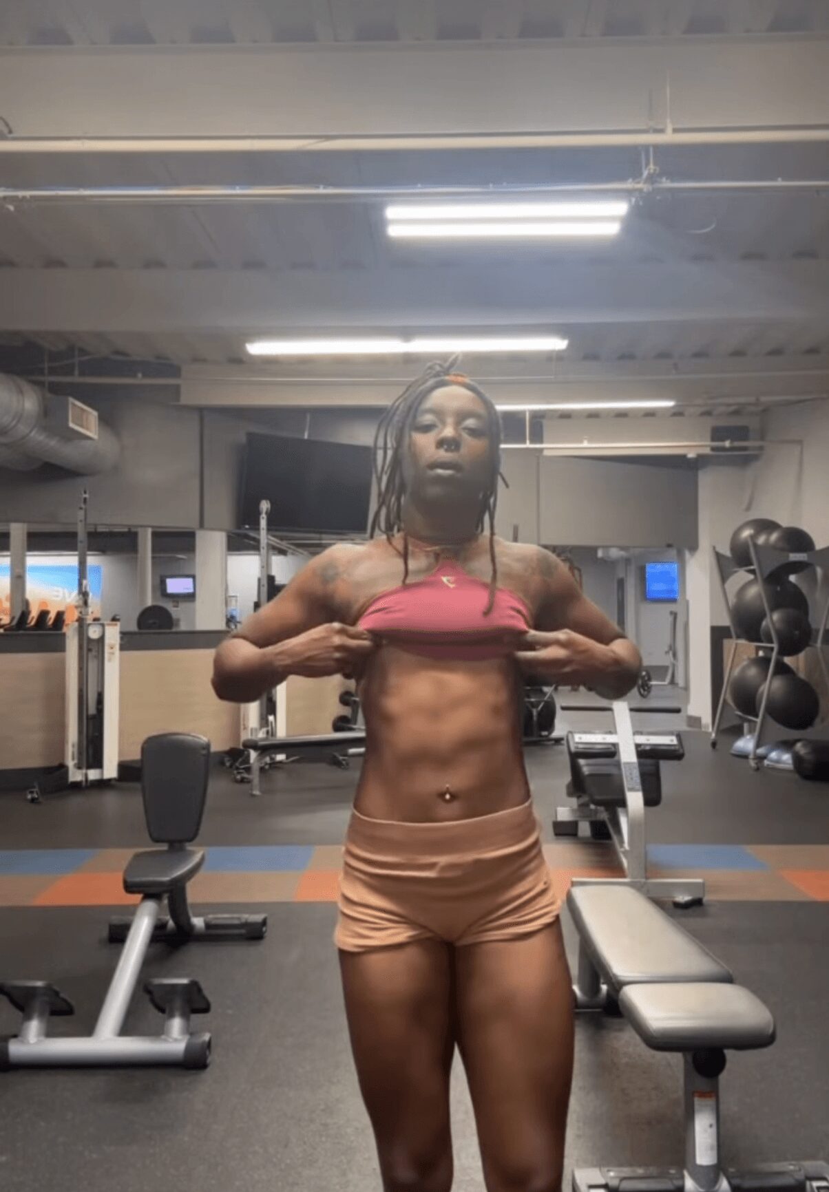 Person standing in gym, lifting a pink top to reveal toned abdomen, wearing beige shorts, with gym equipment in background.