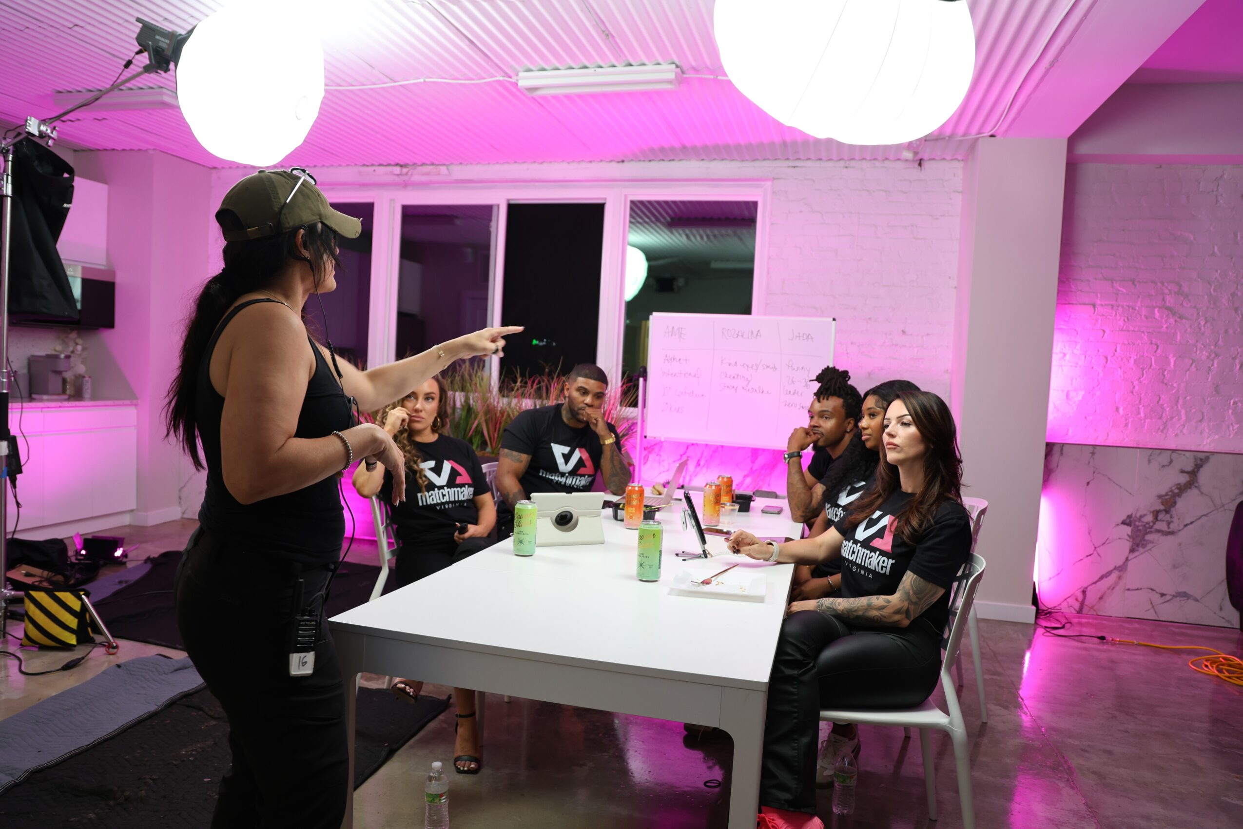 A woman stands and points at a group of five women sitting at a table in a room with pink lighting, white brick walls, and hanging round lights.
