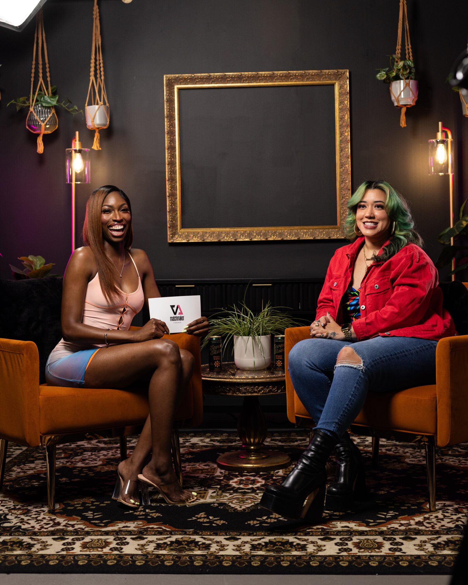 Two women sit on orange chairs in a room with dark walls, hanging plants, and a large empty picture frame behind them.