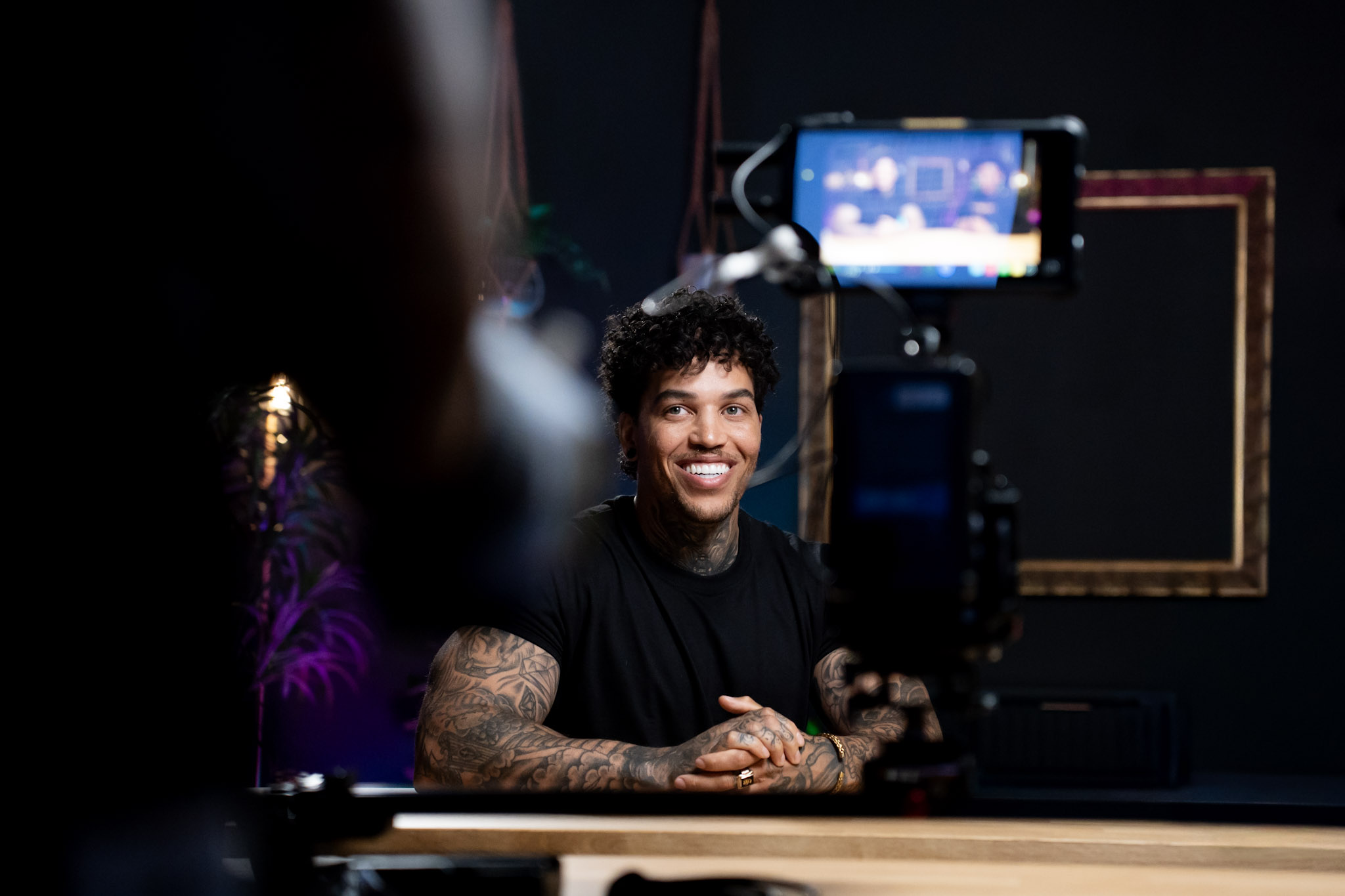 Person with curly hair and tattoos smiling at camera, recording in a dark studio, with a black background and a frame.