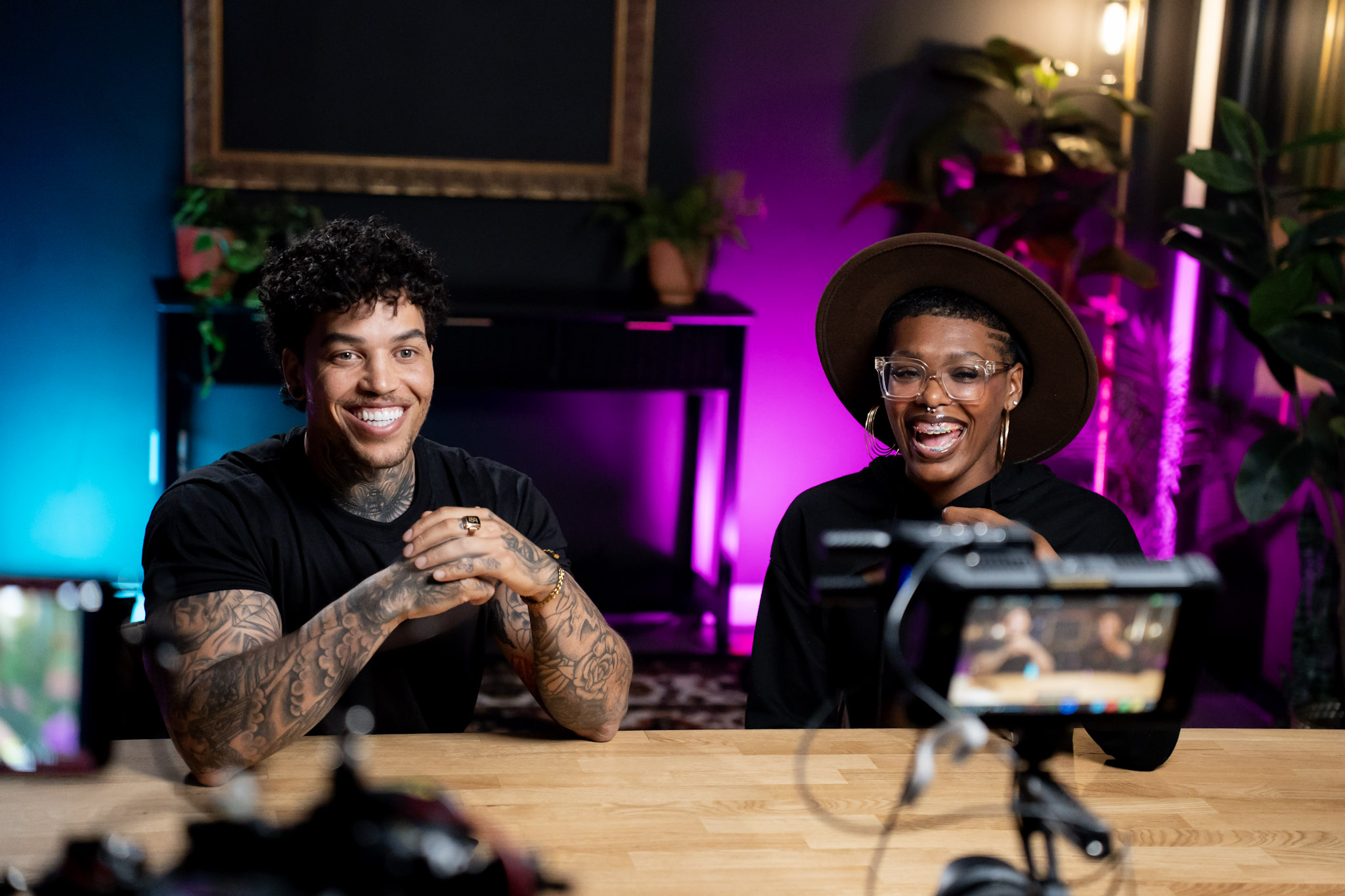 Two people sit at a table, smiling, with a camera recording them, in a colorful room with plants and purple lighting.