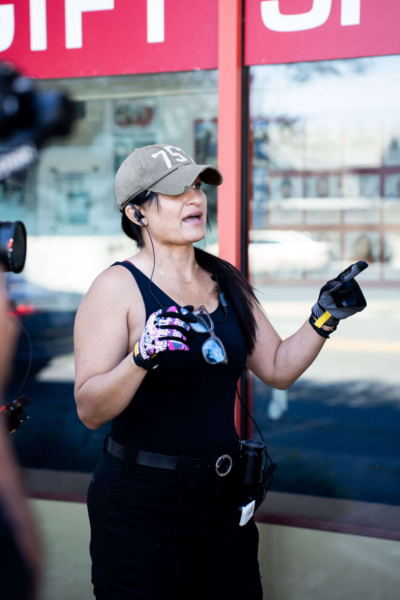 Woman wearing a gray cap, black tank top, and gloves, speaking outdoors with a glass building behind her.