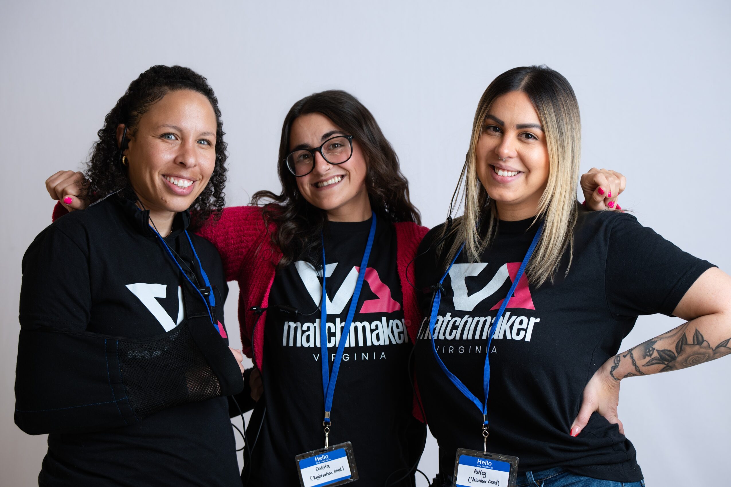 Three women smiling, standing together, wearing black t-shirts with a logo, and blue lanyards, against a light background.