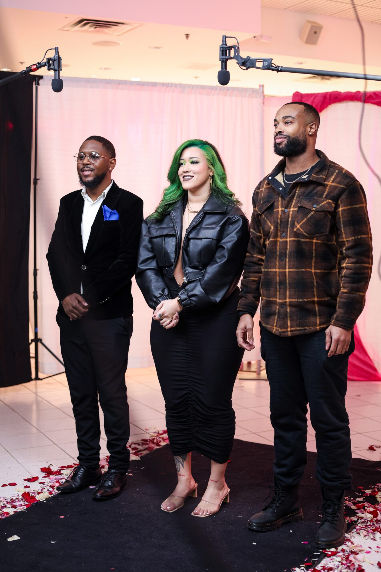 Three people standing on a stage with a pink curtain background, smiling, with a black carpet and red flower petals on the floor.