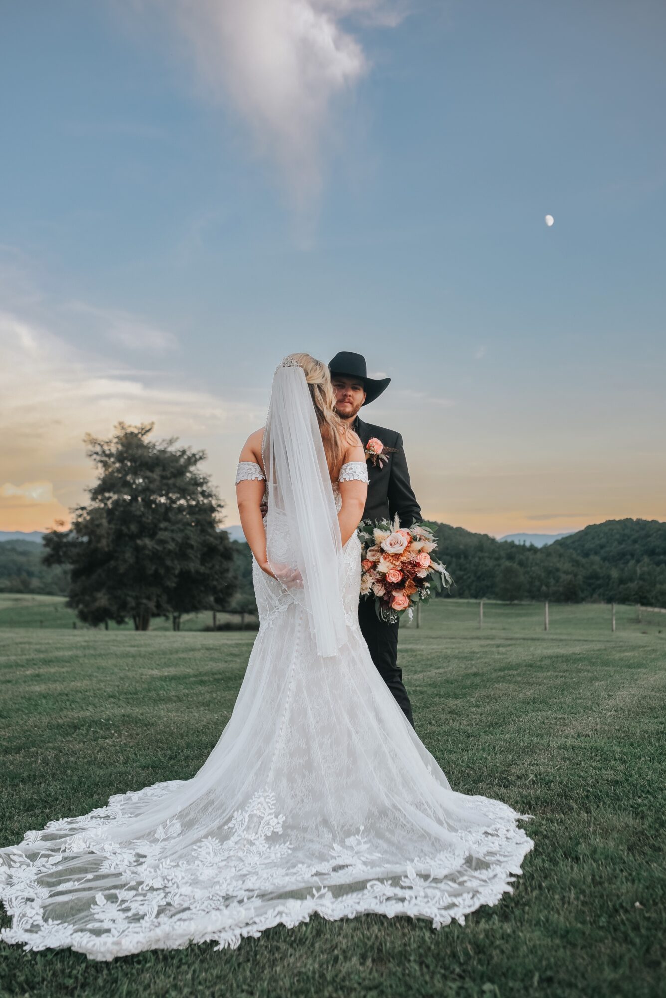 Bride and groom standing outdoors on grass, with trees and hills in the background, during sunset, bride holding a bouquet.